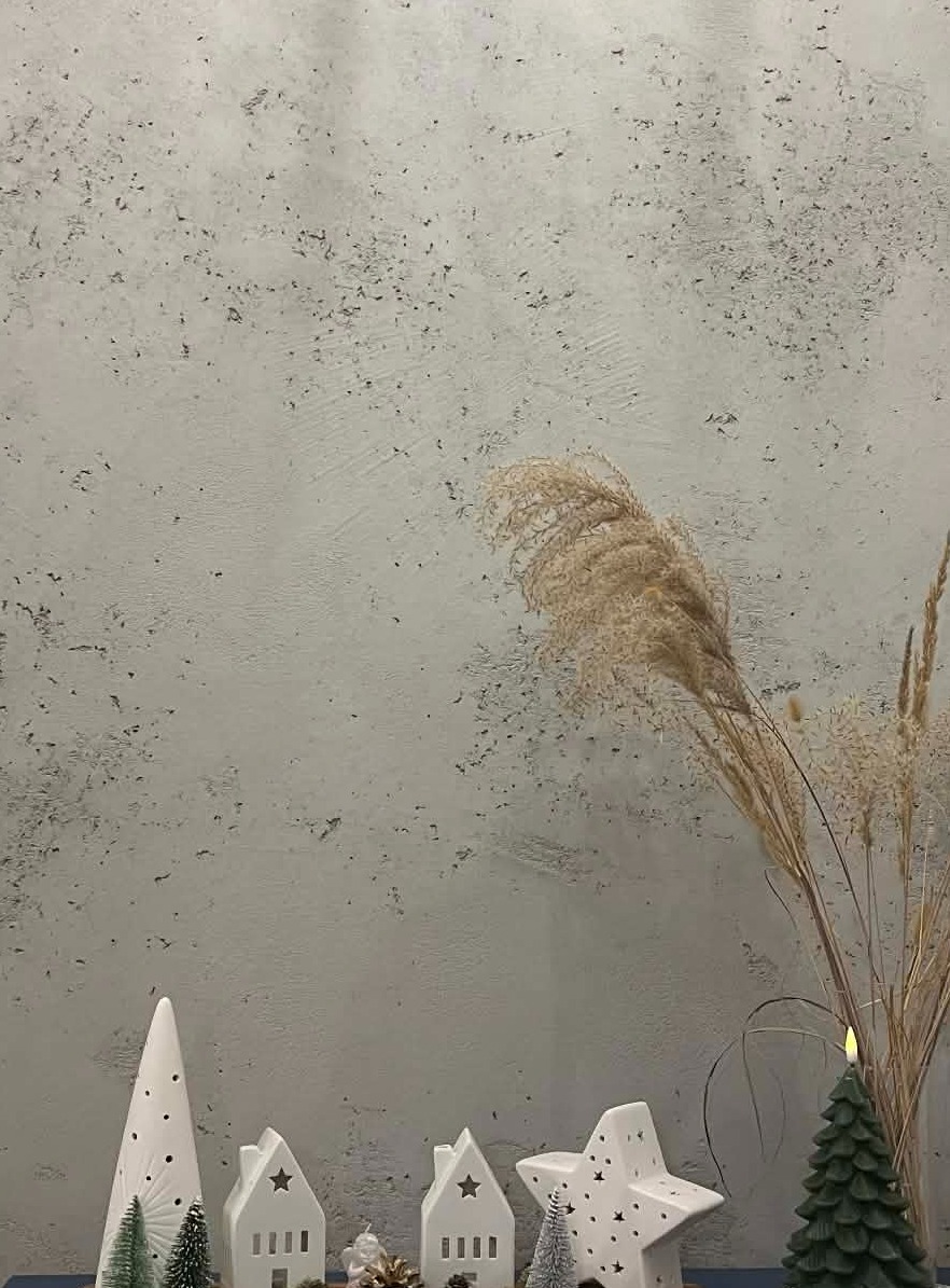 Dekoracyjny tynk w odcieniach szarości z subtelną fakturą, uzupełniony o kompozycję z suszonych traw i ceramicznych ozdób świątecznych w kształcie domków i choinek.