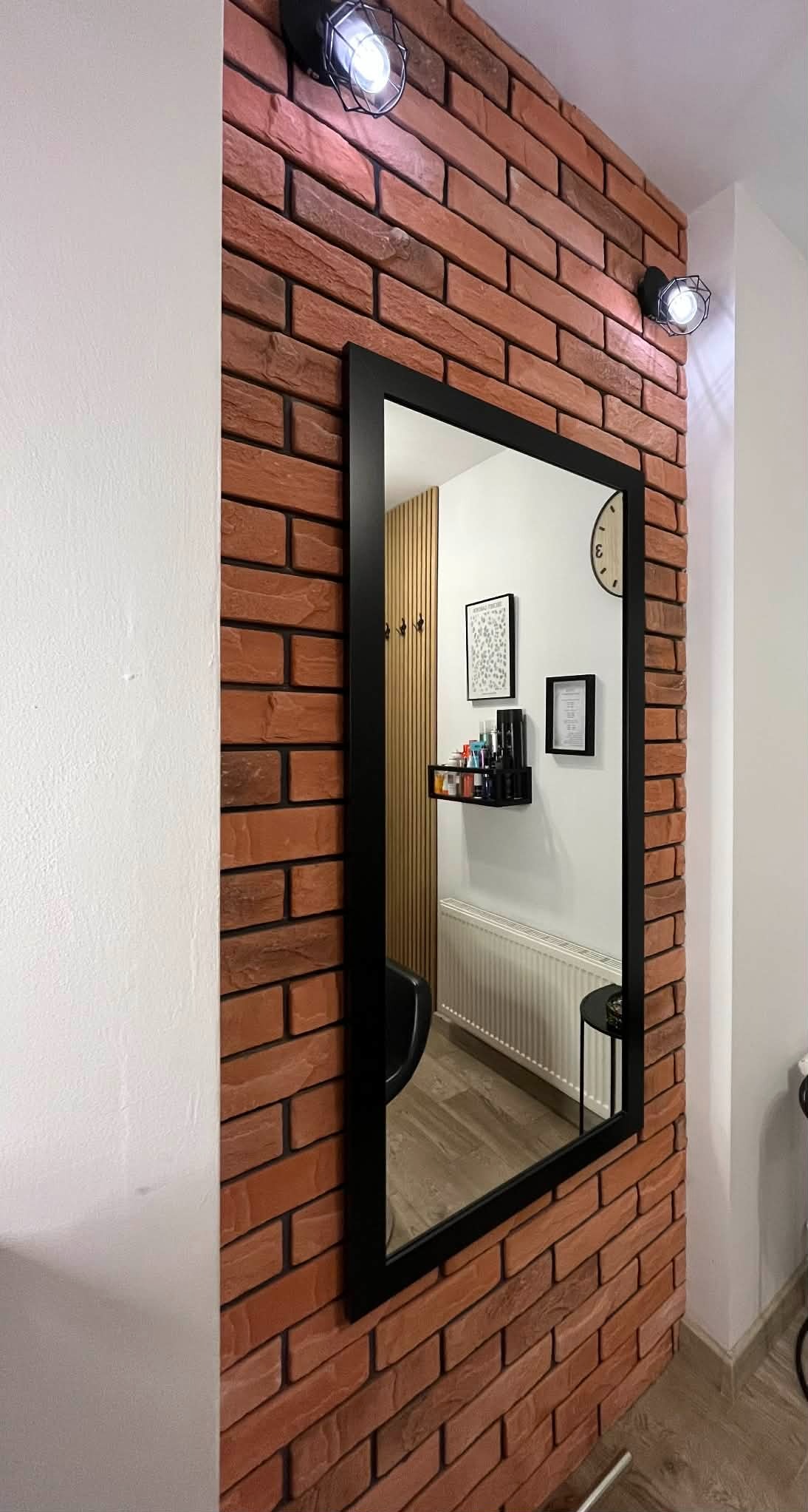 Ściana z cegły klinkierowej z czarnym lustrem, odbijającym fragment wnętrza z lamelami, półką i grzejnikiem. Oświetlenie punktowe na ścianie.