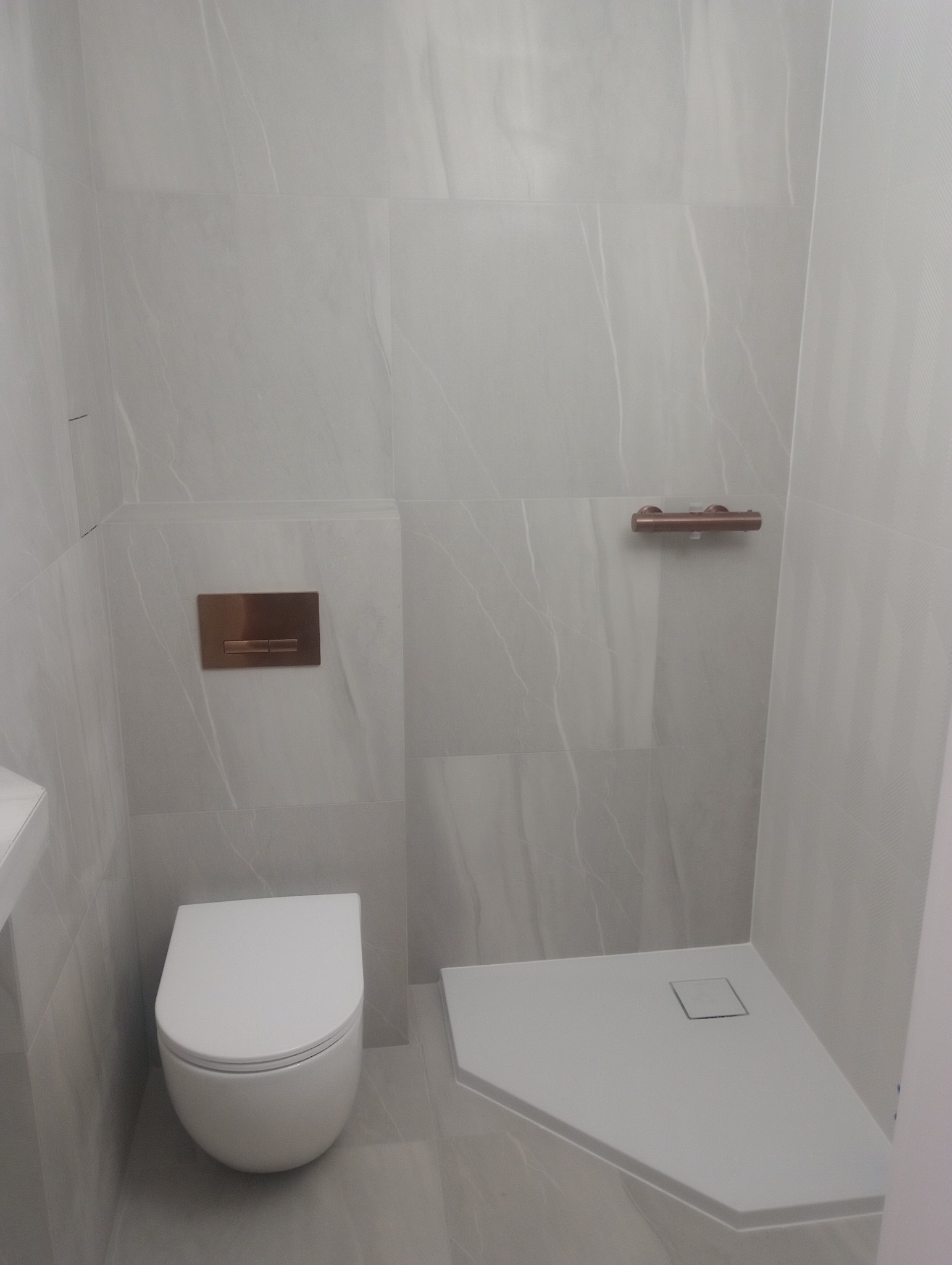 Nowoczesna łazienka z szarymi płytkami imitującymi marmur, wiszącą toaletą i brodzikiem. Miedziane akcenty w postaci przycisku spłuczki i uchwytu na ręcznik.