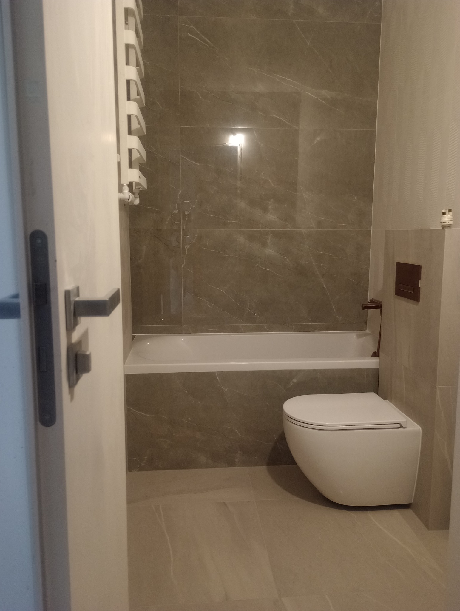 Nowoczesna łazienka z szarymi płytkami imitującymi marmur, wiszącą toaletą i grzejnikiem drabinkowym. Widok zza uchylonych drzwi.