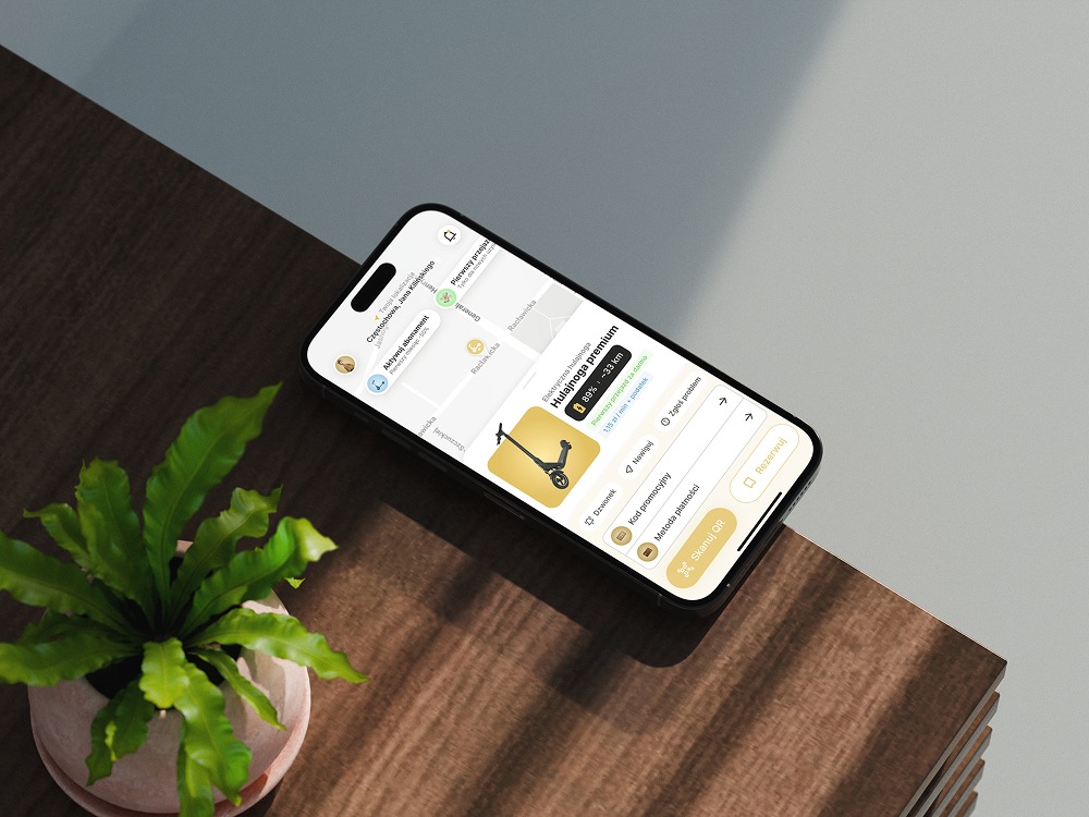 Aplikacja rezerwacji hulajnóg na smartfonie, leżącym na drewnianym blacie obok rośliny doniczkowej. Widoczny interfejs z mapą i ofertą hulajnogi premium.