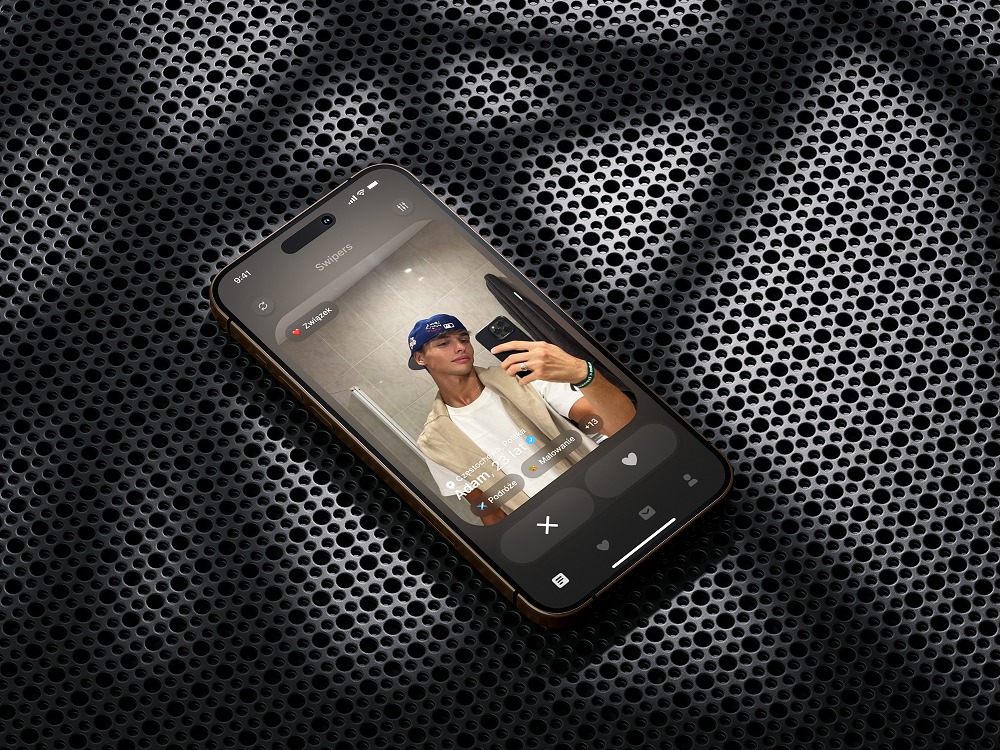 Smartfon z otwartą aplikacją randkową, wyświetlający profil młodego mężczyzny robiącego selfie w lustrze, na tle czarnej, perforowanej powierzchni.