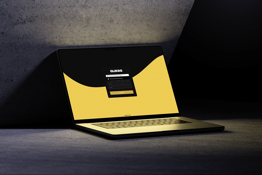 Laptop z ekranem logowania do serwisu Gliddo, na tle betonowej ściany w ciemnym otoczeniu. Minimalistyczny design strony logowania w kolorach czerni i złota.