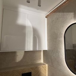 Pro-MB Stolarstwo Mikołaj Barcz - Biała szafka łazienkowa pod sufitem z oświetleniem punktowym, obok lustro z podświetleniem LED. Dekoracyjne wykończenie ściany i drewnopodobna okładzina.