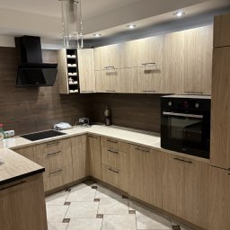 Pro-MB Stolarstwo Mikołaj Barcz - Jasna kuchnia z drewnianymi frontami, czarnym okapem i piekarnikiem. Uwagę przykuwa wzorzysta podłoga i nowoczesna lampa wisząca nad blatem.