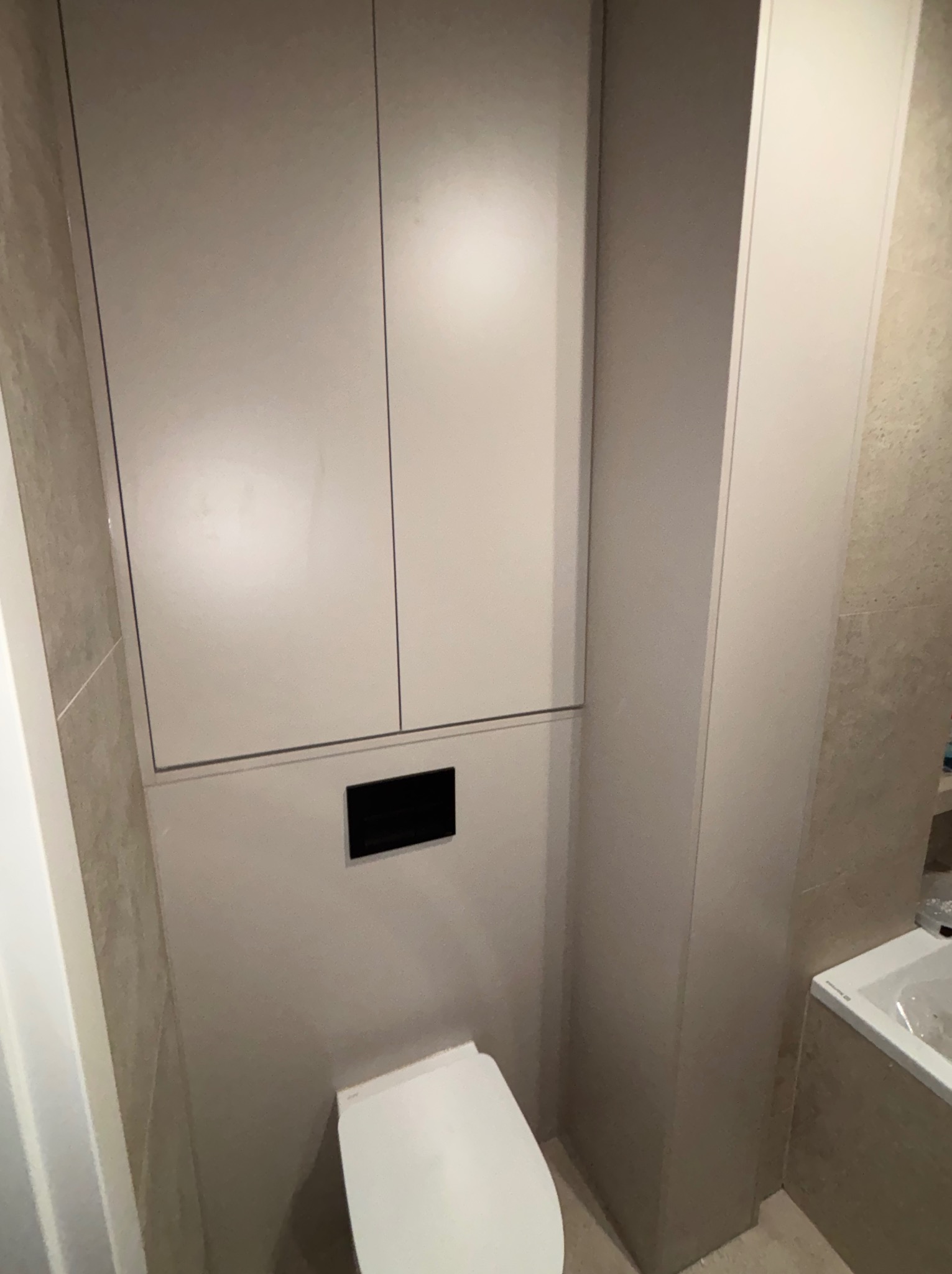 Szafka nad toaletą w łazience w Wejherowie. Zabudowa z szafkami w kolorze beżowym, dopasowana do płytek. Minimalistyczny design, nowoczesne wykończenie.