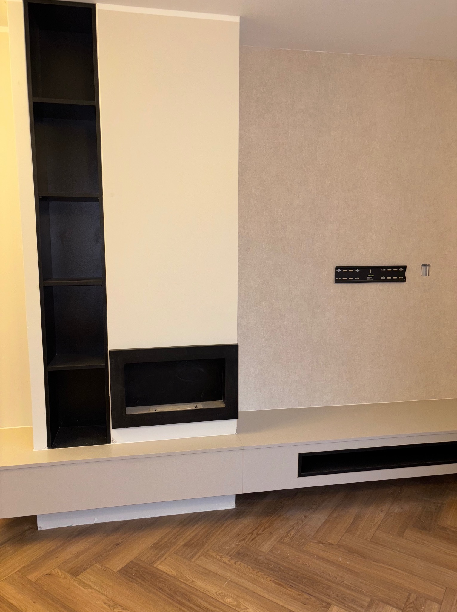 Nowoczesna zabudowa ściany z czarnymi półkami i wnęką na kominek, na tle beżowej ściany z uchwytem TV. Parkiet w jodełkę na podłodze.