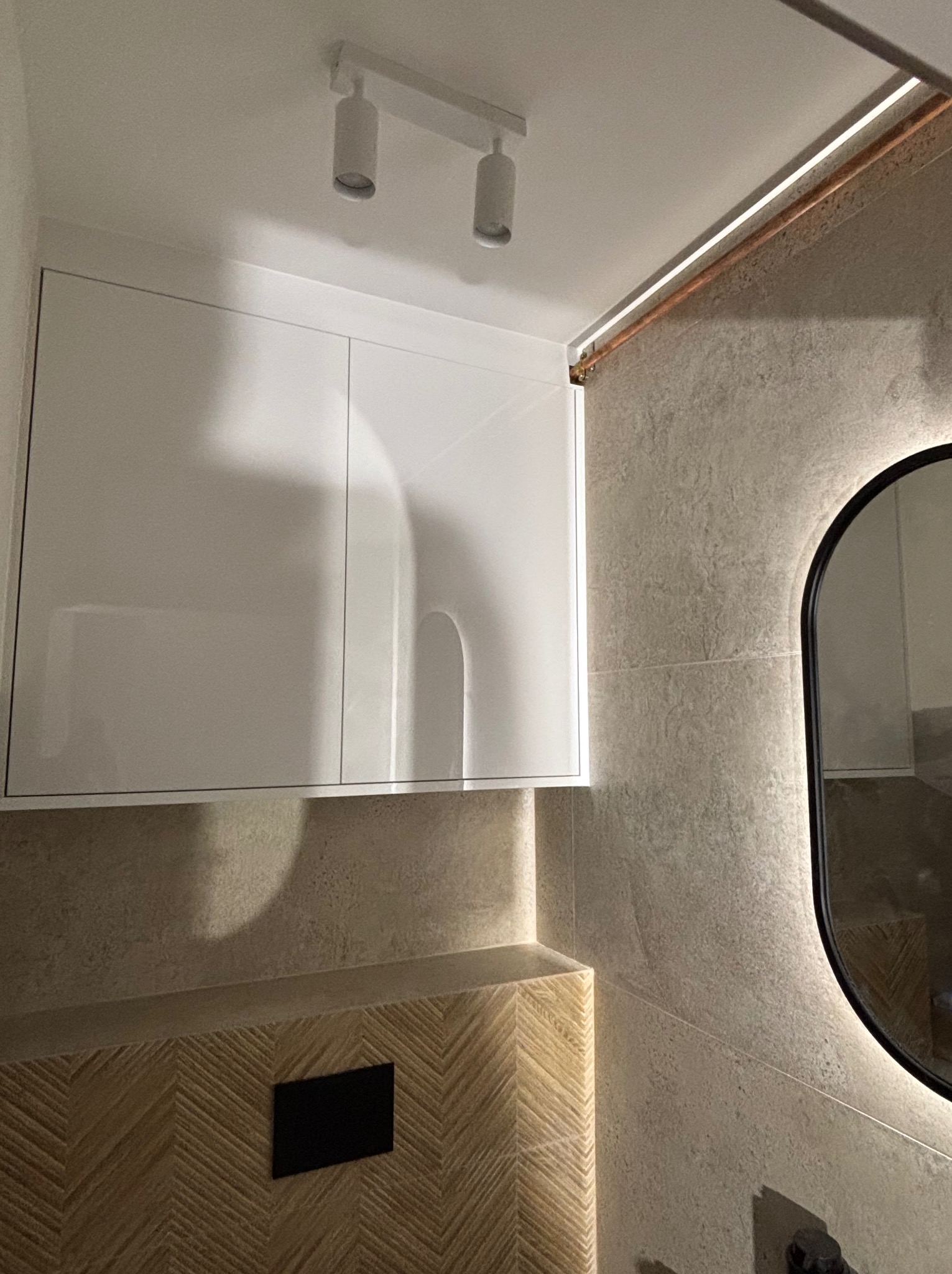 Biała szafka łazienkowa pod sufitem z oświetleniem punktowym, obok lustro z podświetleniem LED. Dekoracyjne wykończenie ściany i drewnopodobna okładzina.