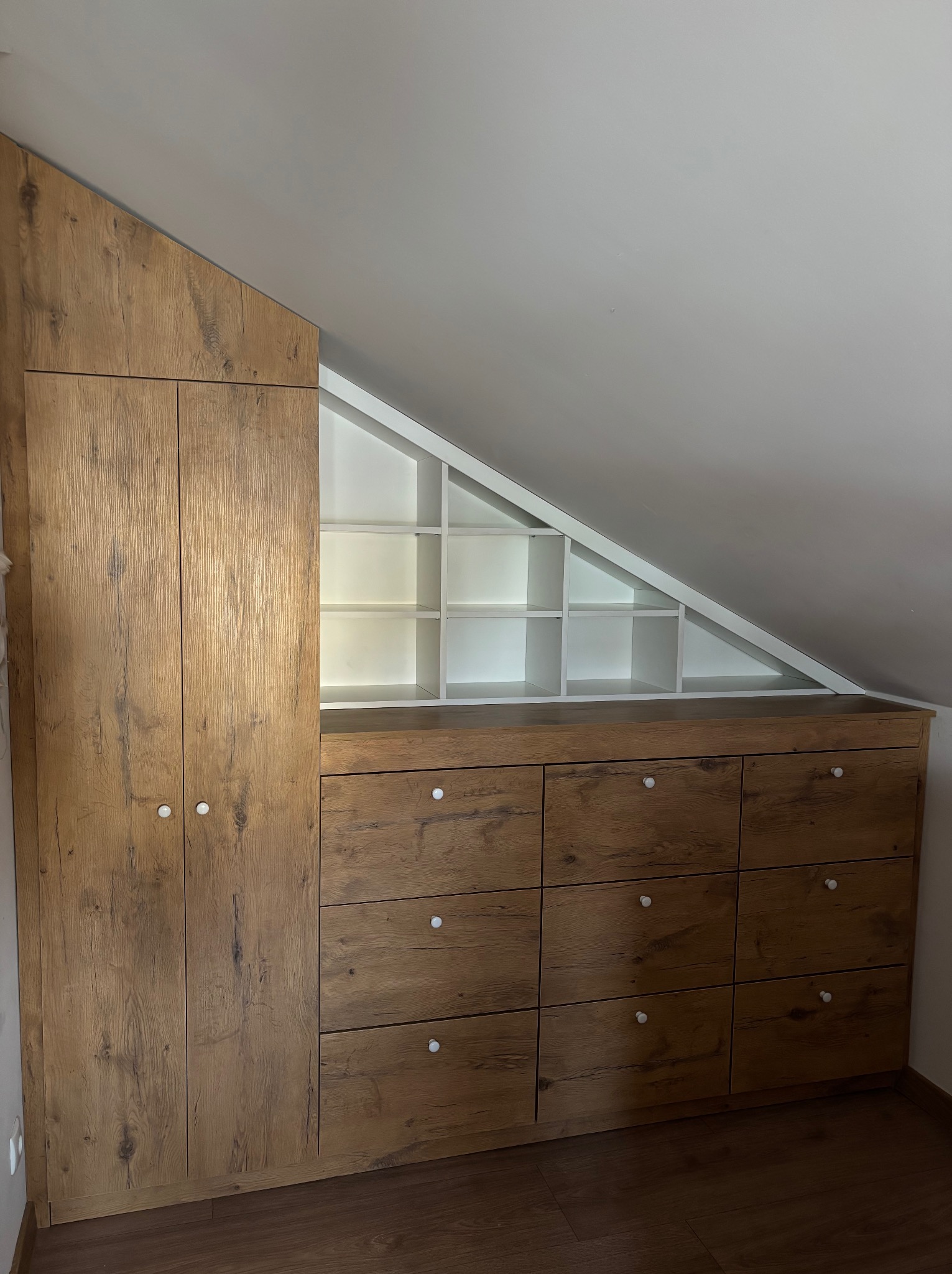 Drewniana szafa z szufladami i półkami w skosie poddasza, białe wnętrze półek, okrągłe uchwyty, podłoga z paneli, dopasowanie do przestrzeni.