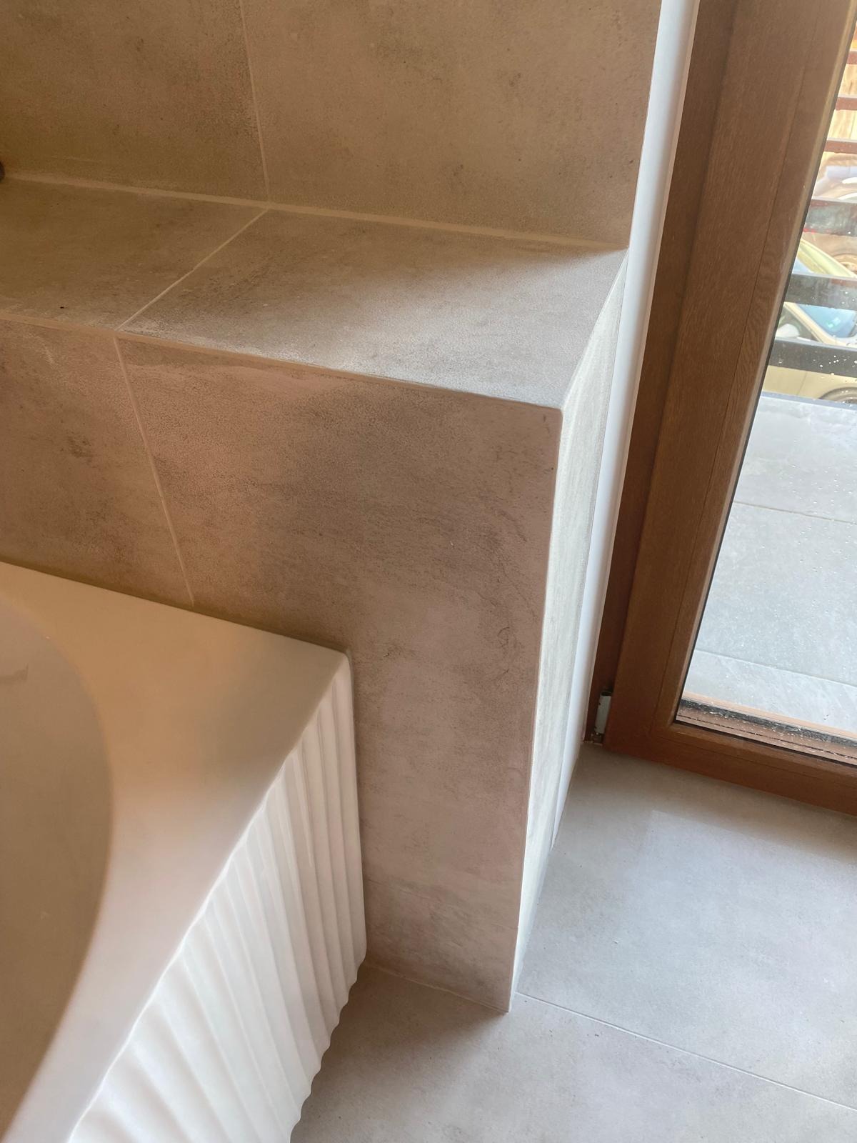 Wykończona łazienka: beżowe płytki na ścianie i podłodze, fragment wanny z dekoracyjnym wykończeniem, drewniane obramowanie okna. Minimalistyczny design.