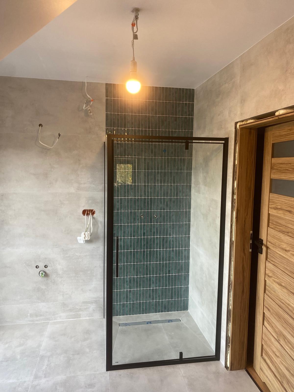 Nowoczesna kabina prysznicowa z czarną ramą i zielonymi płytkami w łazience z szarymi płytkami. Widoczne niedokończone prace elektryczne.