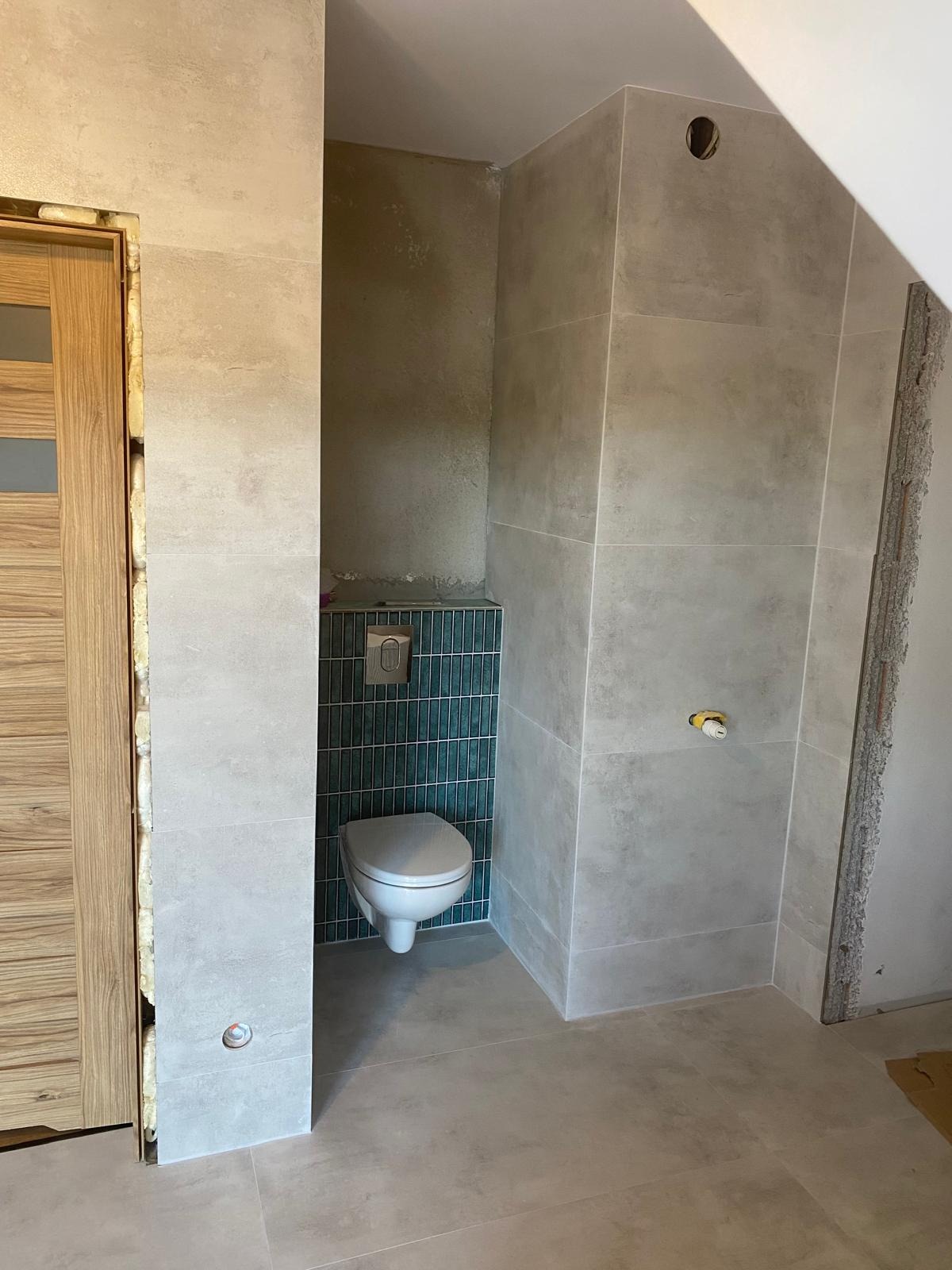 Wykończona łazienka w odcieniach beżu i szarości z akcentem turkusowej mozaiki za toaletą. Widoczne płytki na podłodze i ścianach, częściowo wykończone obramowanie drzwi.
