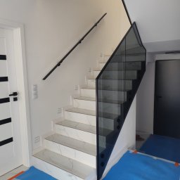 SD Budownictwo - Schody z marmurowymi stopniami i szklaną balustradą z czarnymi detalami, widok z dołu. Nowoczesny design, minimalistyczne wykończenie.