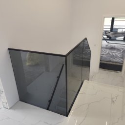 SD Budownictwo - Szklana balustrada schodowa z czarną ramą, widok z góry. Białe marmurowe schody i podłoga. W tle widoczny pokój z hamakiem.