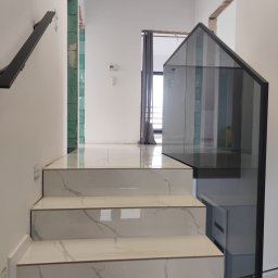 SD Budownictwo - Szklana balustrada schodowa z czarną ramą i marmurowymi schodami, widok z dołu. Minimalistyczny design, jasne wnętrze, częściowo widoczny fragment poręczy.