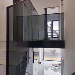 SD Budownictwo - Szklana balustrada schodowa z czarnymi elementami mocującymi, widok z góry na parter, nowoczesny design, jasne wnętrze z dużymi oknami i widokiem na zewnątrz.