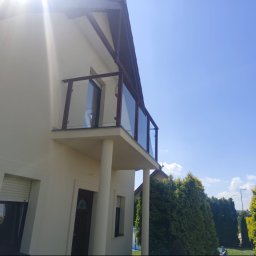 SD Budownictwo - Balkon na piętrze nowoczesnego domu z beżową elewacją, wyposażony w szklaną barierę z ciemnobrązowymi słupkami. Słoneczny dzień, błękitne niebo i zielone tuje w tle.
