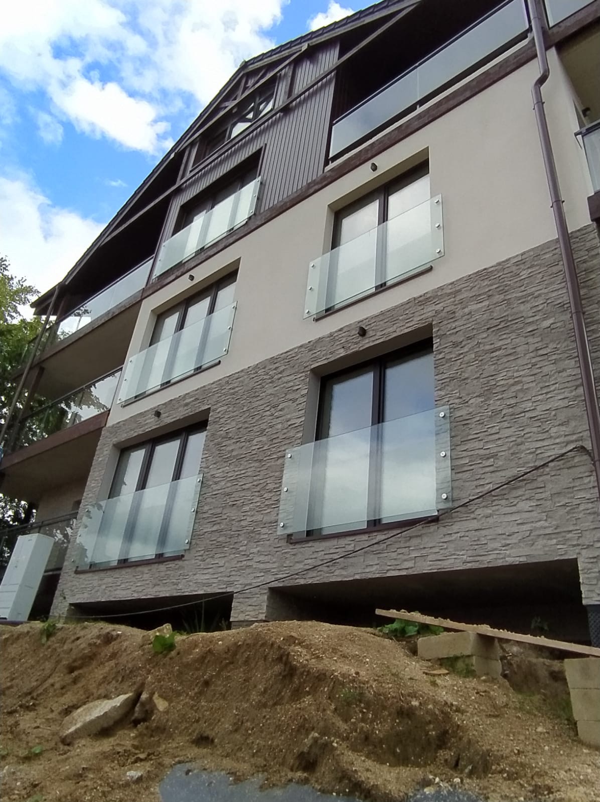 Nowoczesny budynek z balkonami zabezpieczonymi szkłem, widok z dołu na tle błękitnego nieba. Elewacja z kamienia i drewna. Uwagę przykuwają szklane balustrady.