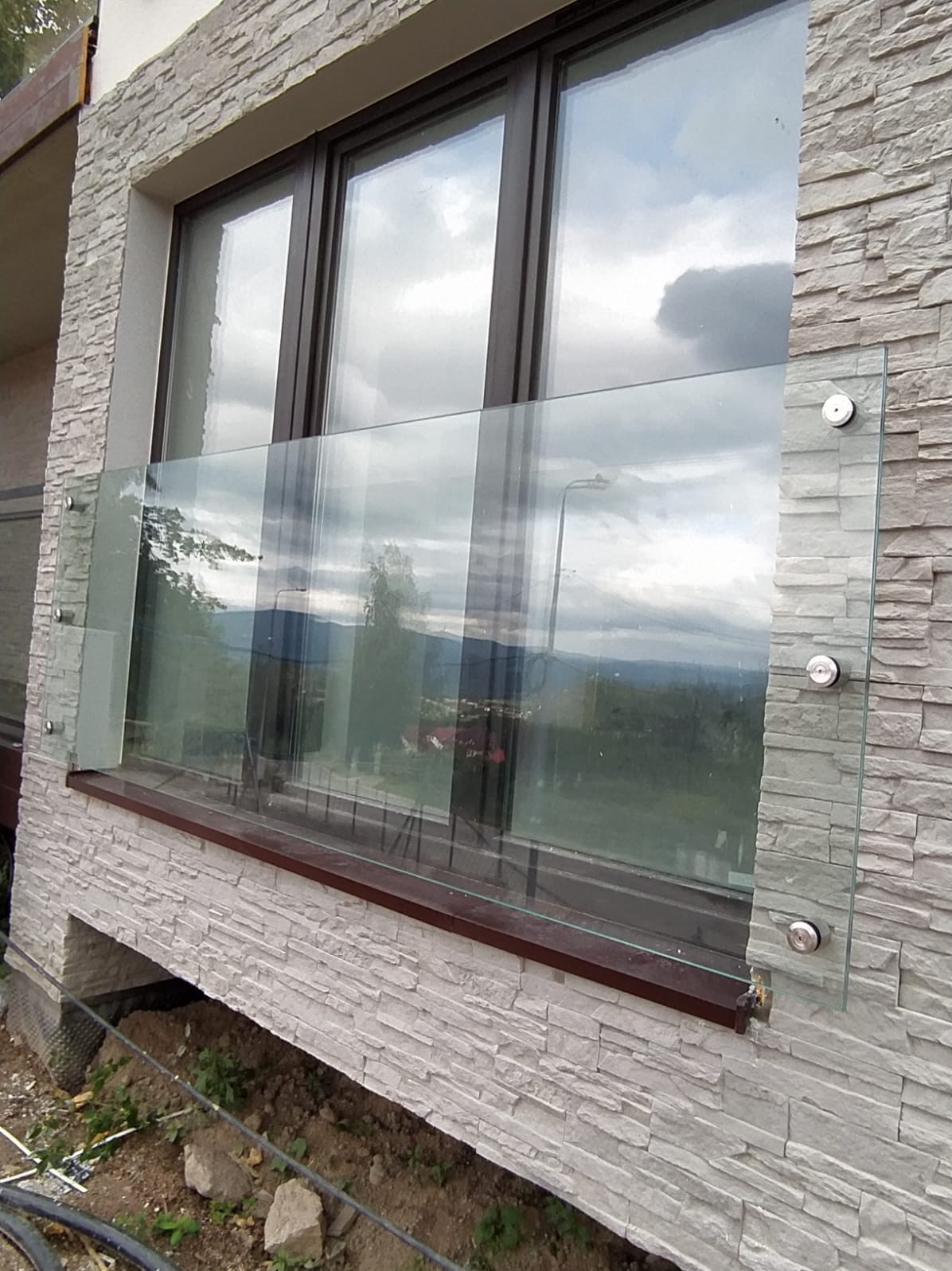 Szklana balustrada balkonowa z widokiem na góry, mocowana na ozdobnej elewacji z kamienia. Widoczne metalowe uchwyty mocujące szkło. Ujęcie z dołu.