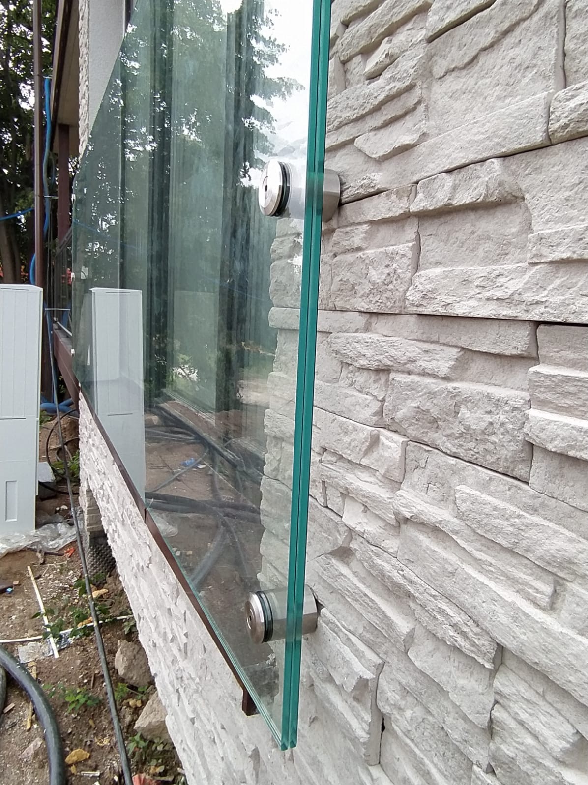 Szklana balustrada mocowana do ściany z kamienia dekoracyjnego za pomocą metalowych uchwytów. Widoczne elementy konstrukcyjne i otoczenie zewnętrzne.