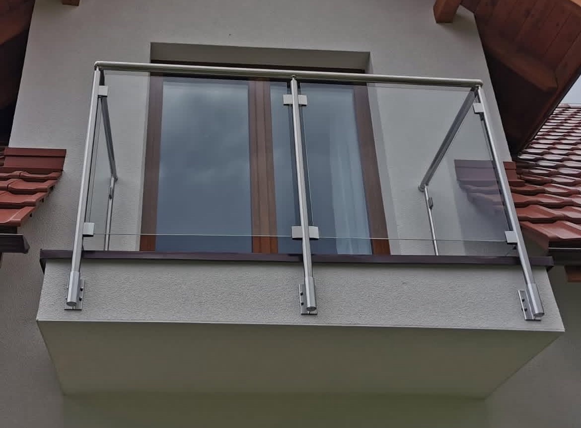 Szklana balustrada balkonowa z elementami ze stali nierdzewnej, zamontowana na balkonie budynku z brązowymi oknami i czerwoną dachówką w tle.