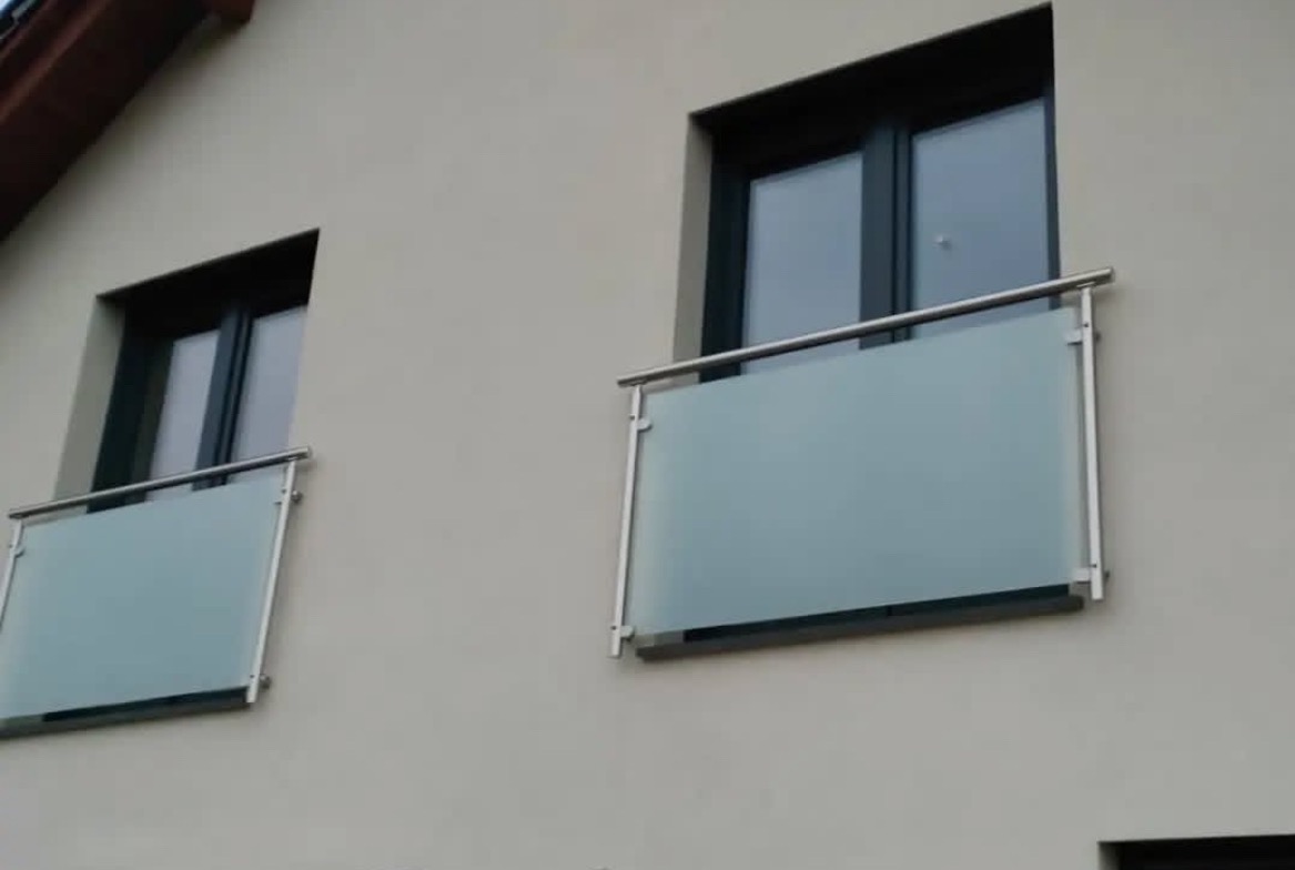 Szklane balustrady balkonowe z nierdzewnymi poręczami, zamontowane przy oknach na elewacji budynku. Minimalistyczny design, matowe szkło.