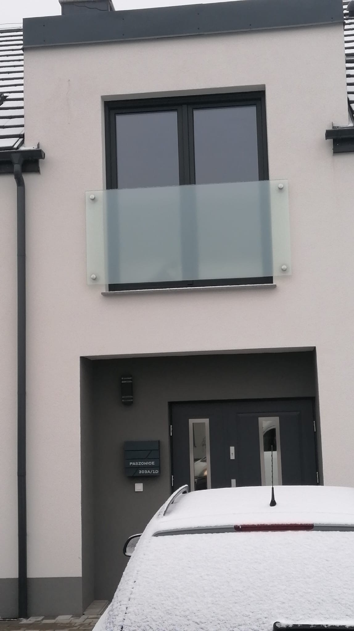 Szklana balustrada balkonowa z matowego szkła, zamontowana na nowoczesnym budynku w zimowej scenerii. Widoczny fragment ośnieżonego samochodu.