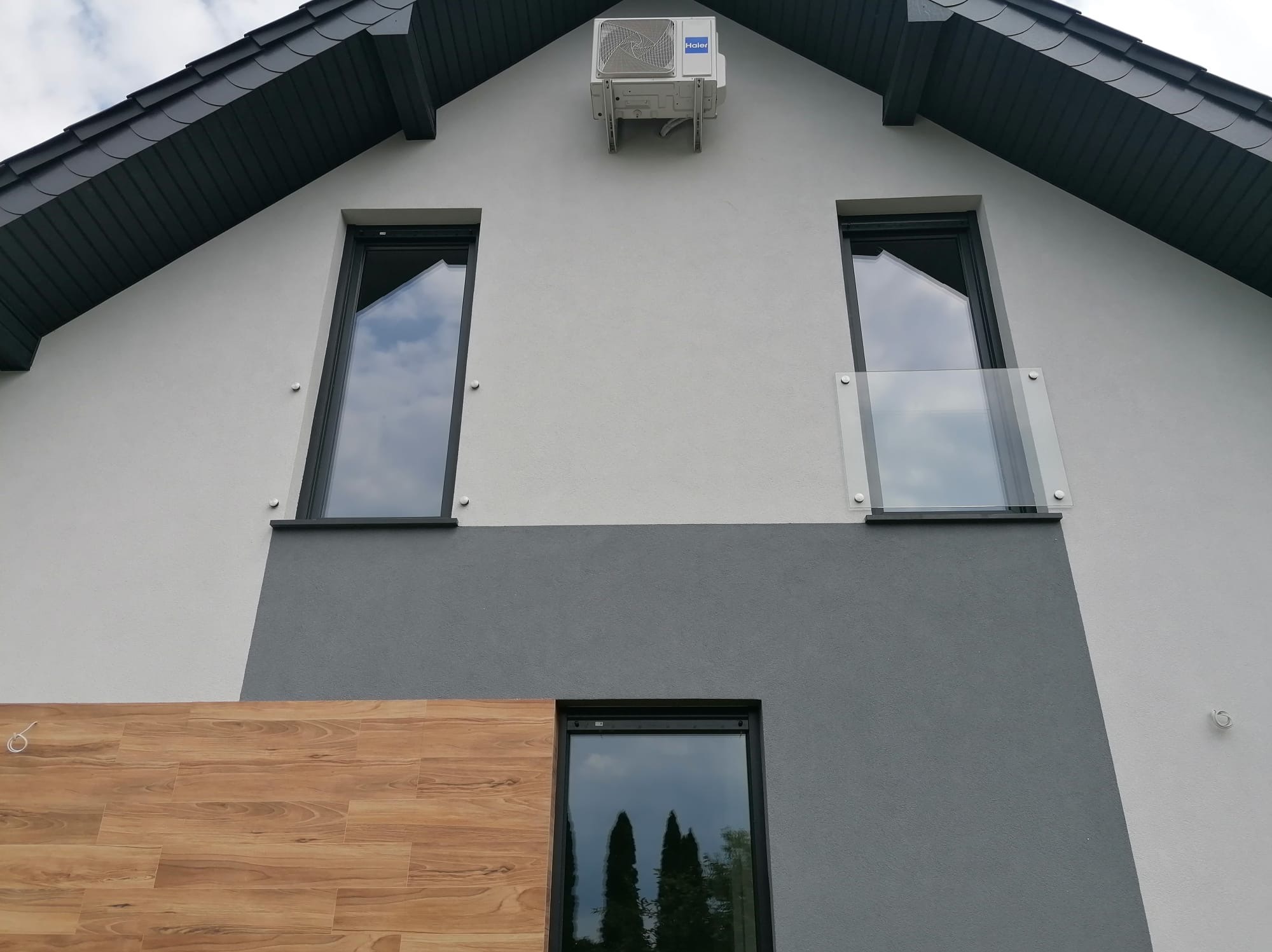 Nowoczesna elewacja domu z oknami i szklaną balustradą przy oknie na piętrze. Widoczny fragment drewnianej okładziny i klimatyzator na szczycie dachu.