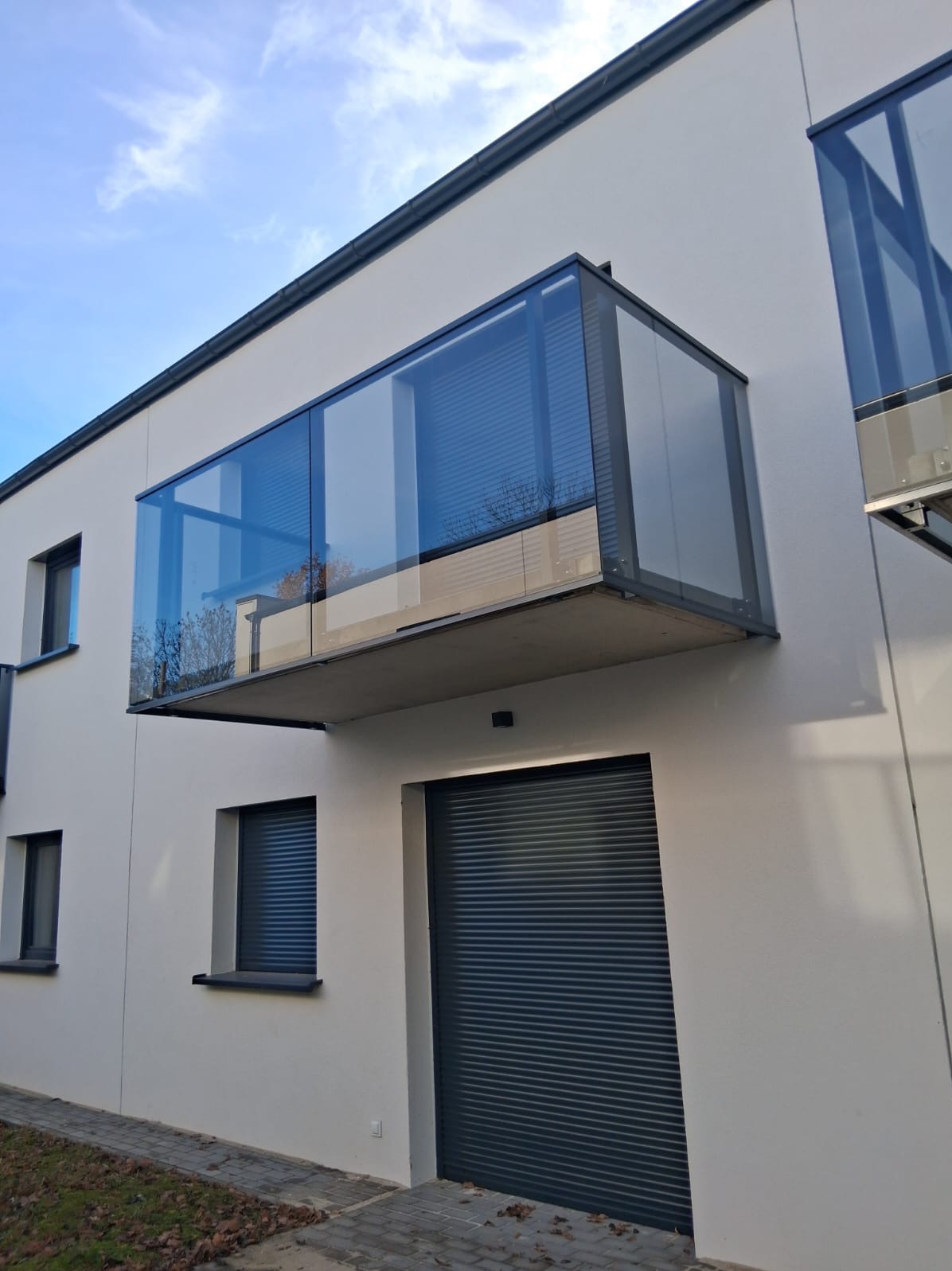 Nowoczesny balkon z szklaną balustradą, widok z dołu, szare ramy, minimalistyczny design, biała elewacja budynku, rolety zewnętrzne w oknach, błękitne niebo.