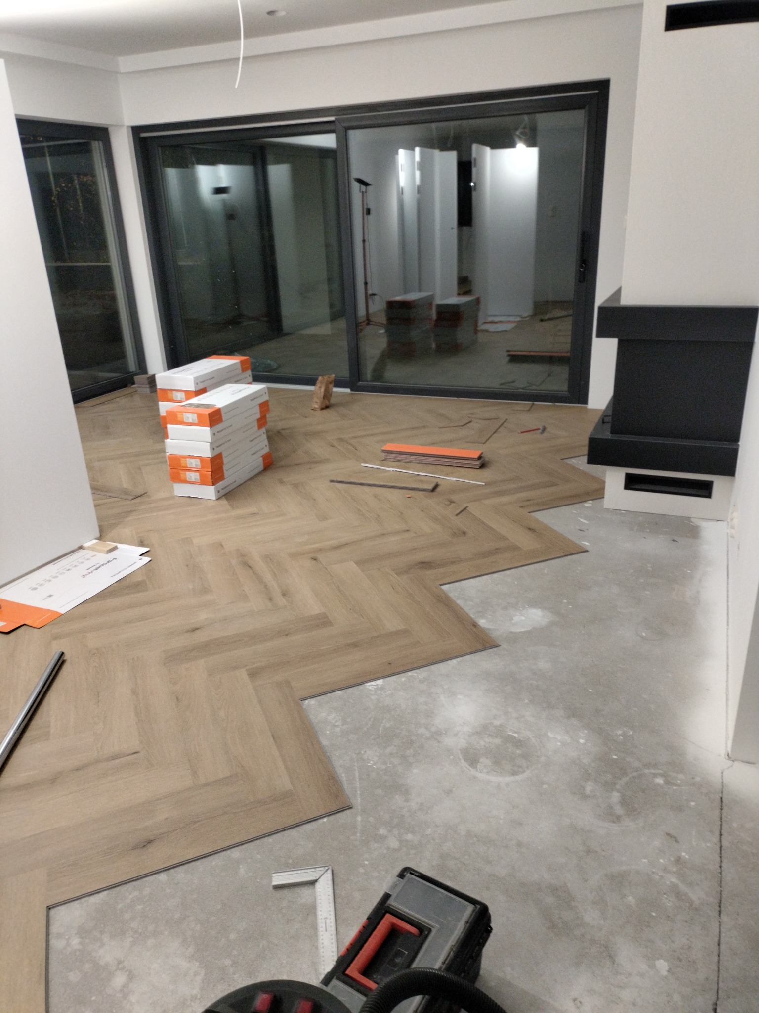 Układanie paneli w jodełkę: widok na częściowo wykończoną podłogę, paczki paneli i narzędzia. W tle przesuwne drzwi balkonowe i fragment kominka.