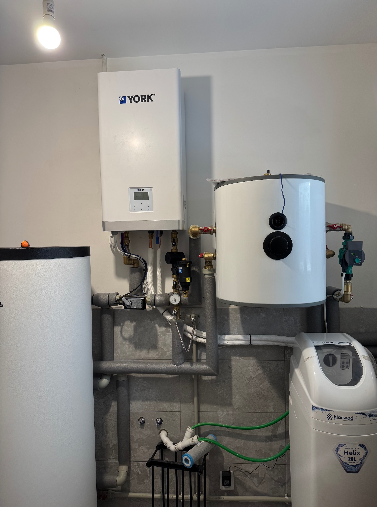 Biały piec gazowy York, zbiornik na wodę i zmiękczacz wody Klarwod Helix 20L, połączone rurami w kotłowni z szarymi płytkami. Widoczne zawory i manometry.