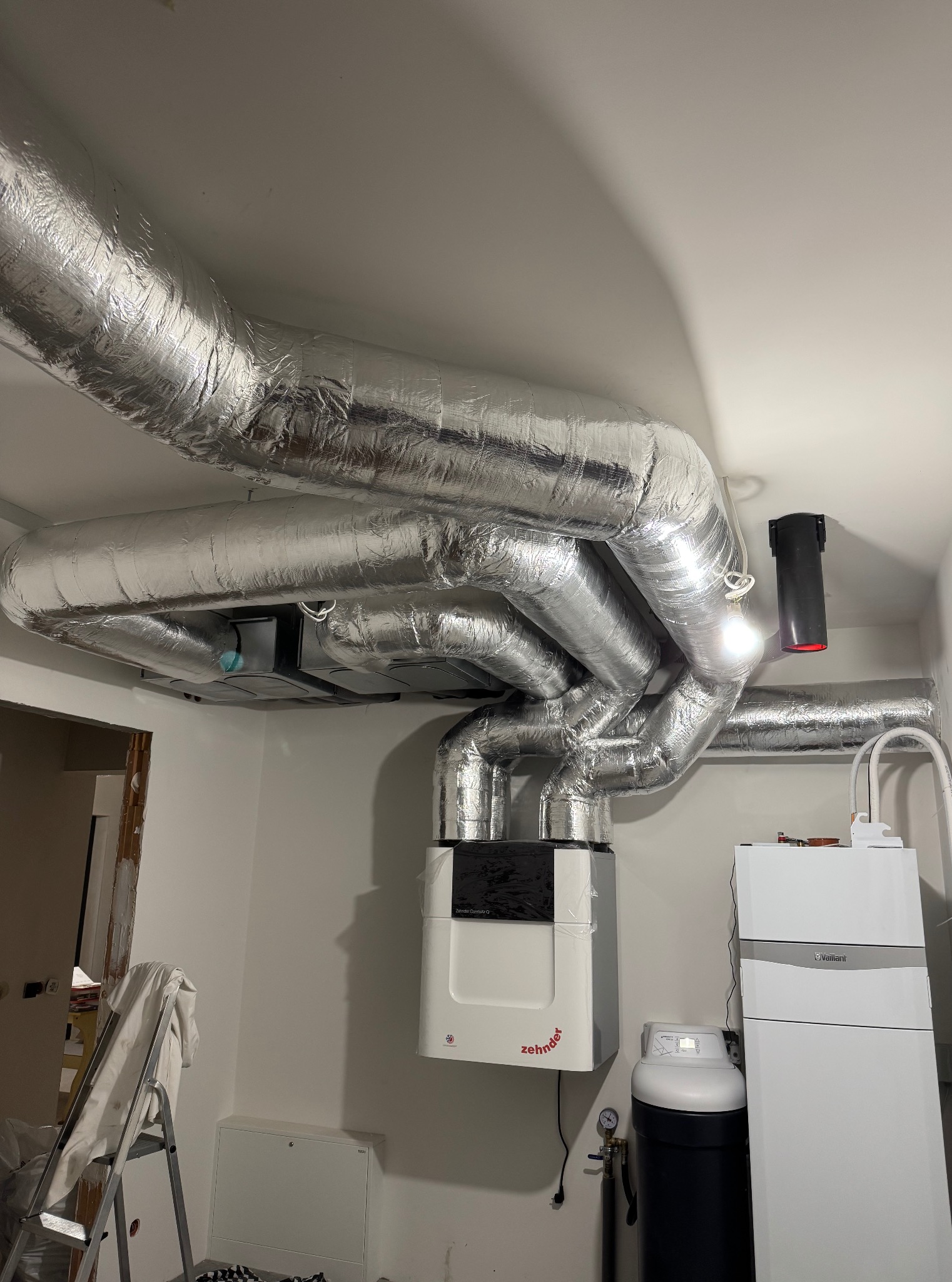 Złożony system wentylacyjny z rurami w srebrnej izolacji, zamontowany pod sufitem w pomieszczeniu z białymi ścianami. Widoczny piec Zehnder i elementy instalacji Vaillant.