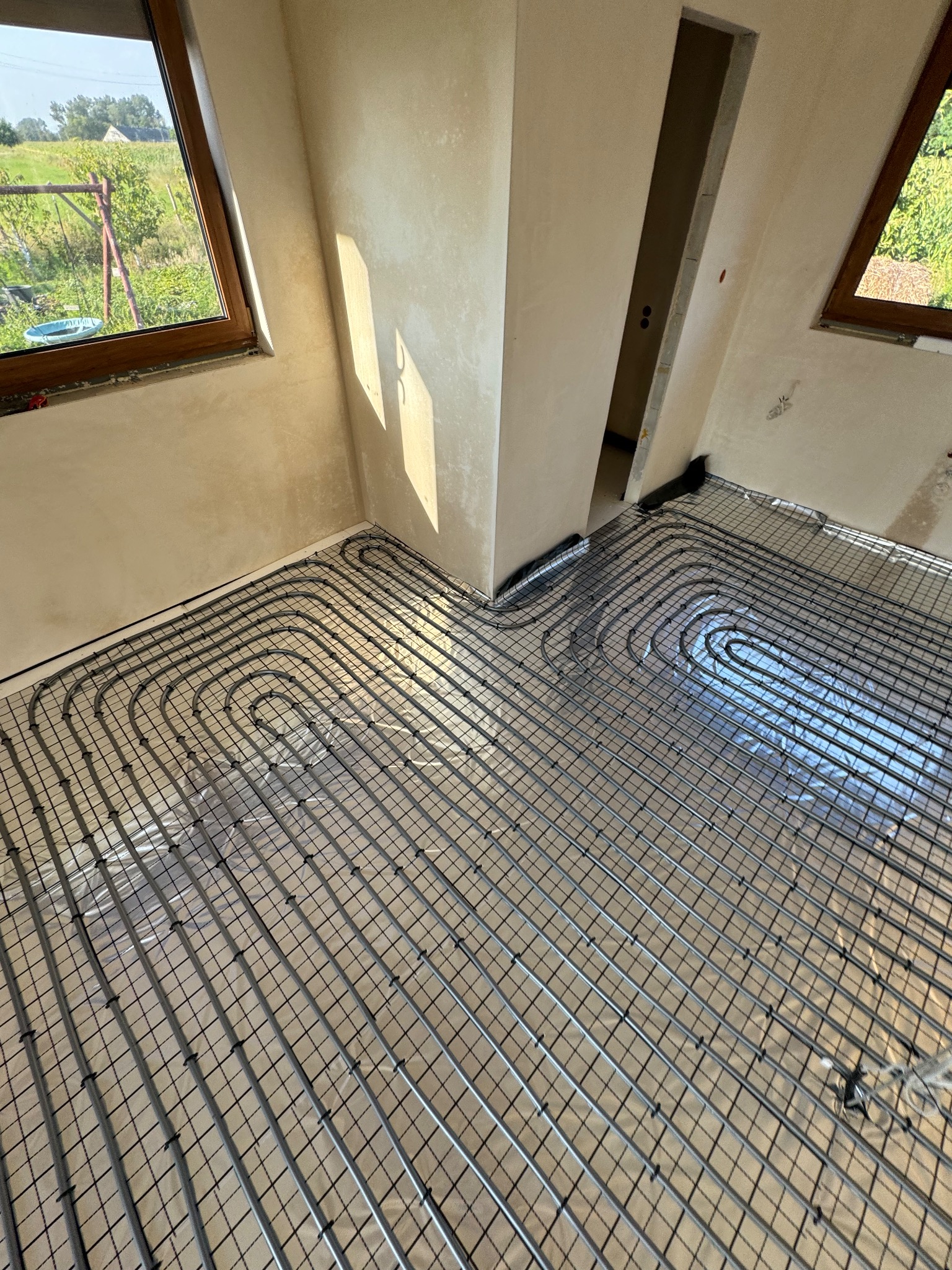 Ułożona instalacja ogrzewania podłogowego na folii izolacyjnej w jasnym pomieszczeniu z oknami. Widoczne rury w kształcie pętli, przygotowane do zalania wylewką.