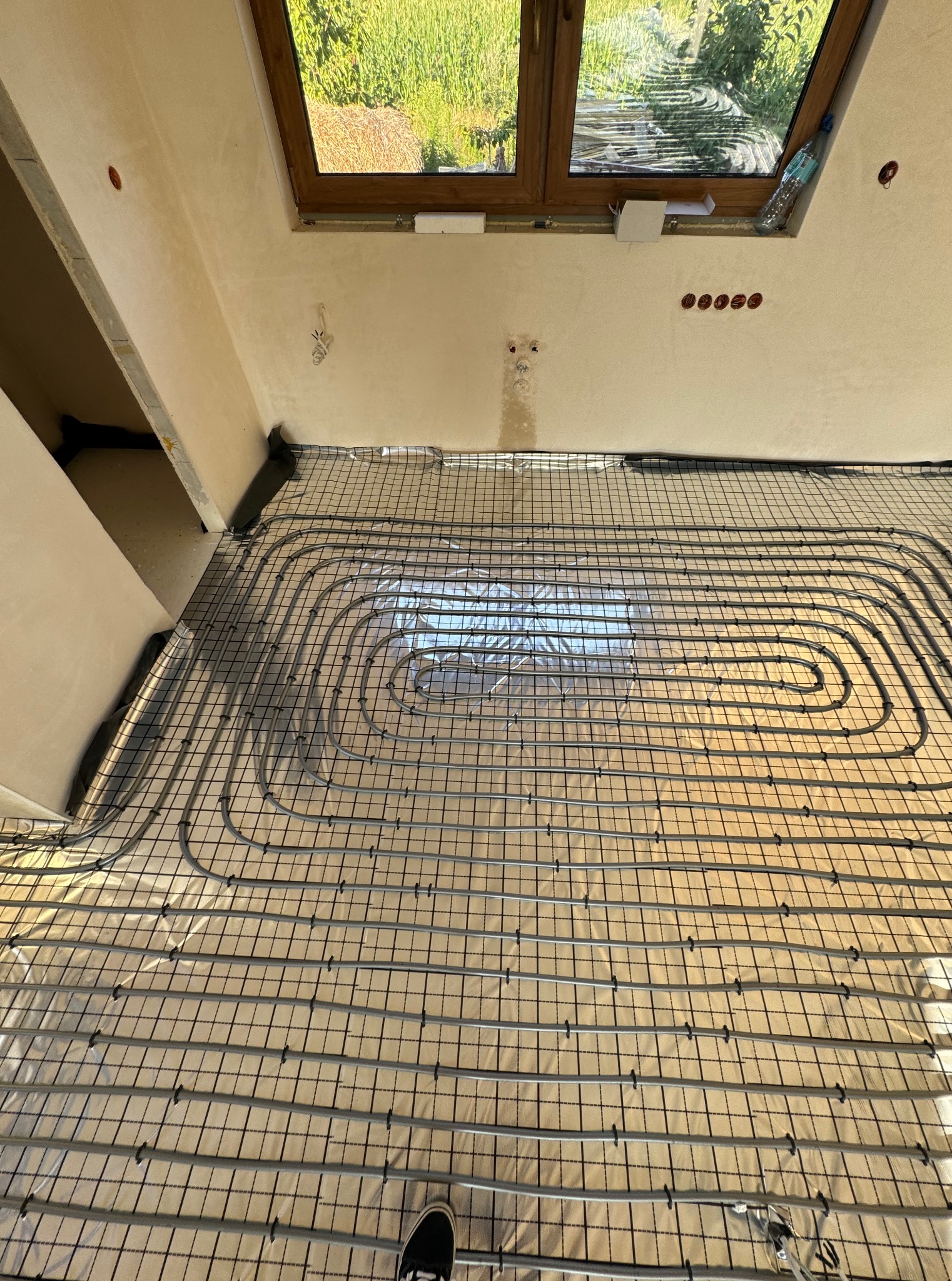 Szare rury ogrzewania podłogowego ułożone na folii izolacyjnej z siatką, widok z góry, fragment buta w rogu. Okno w tle, ściany w stanie surowym.