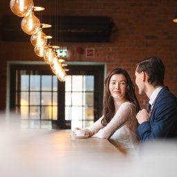 SHOTMAX - Uśmiechnięta panna młoda w koronkowej sukni siedzi przy barze z panem młodym w granatowym garniturze, pod dekoracyjnymi żarówkami w ciepłym świetle.