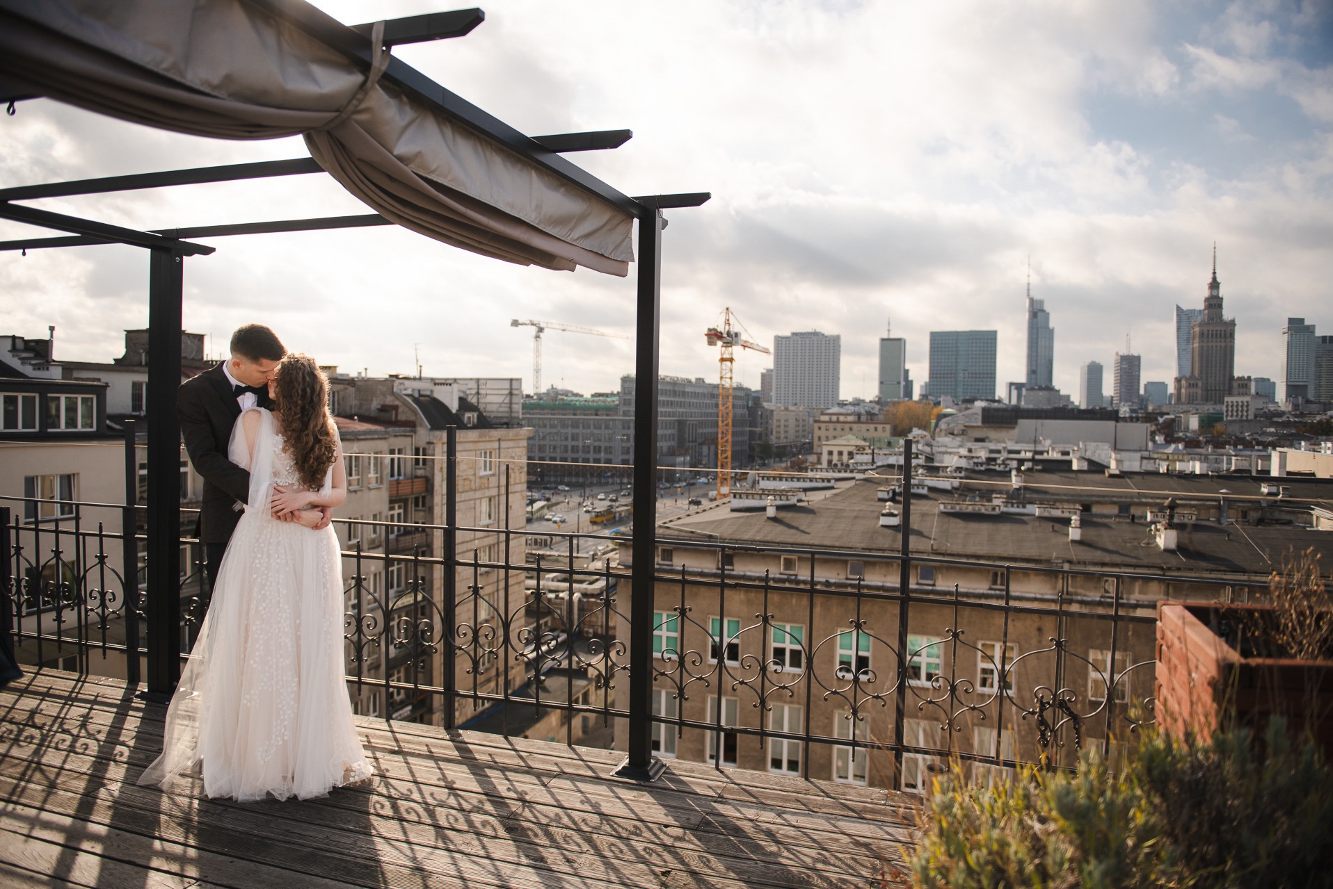 Para młoda w objęciach na tle panoramy miasta z Pałacem Kultury, sfotografowana z dachu budynku z pergolą. Romantyczna sesja w miejskiej scenerii.