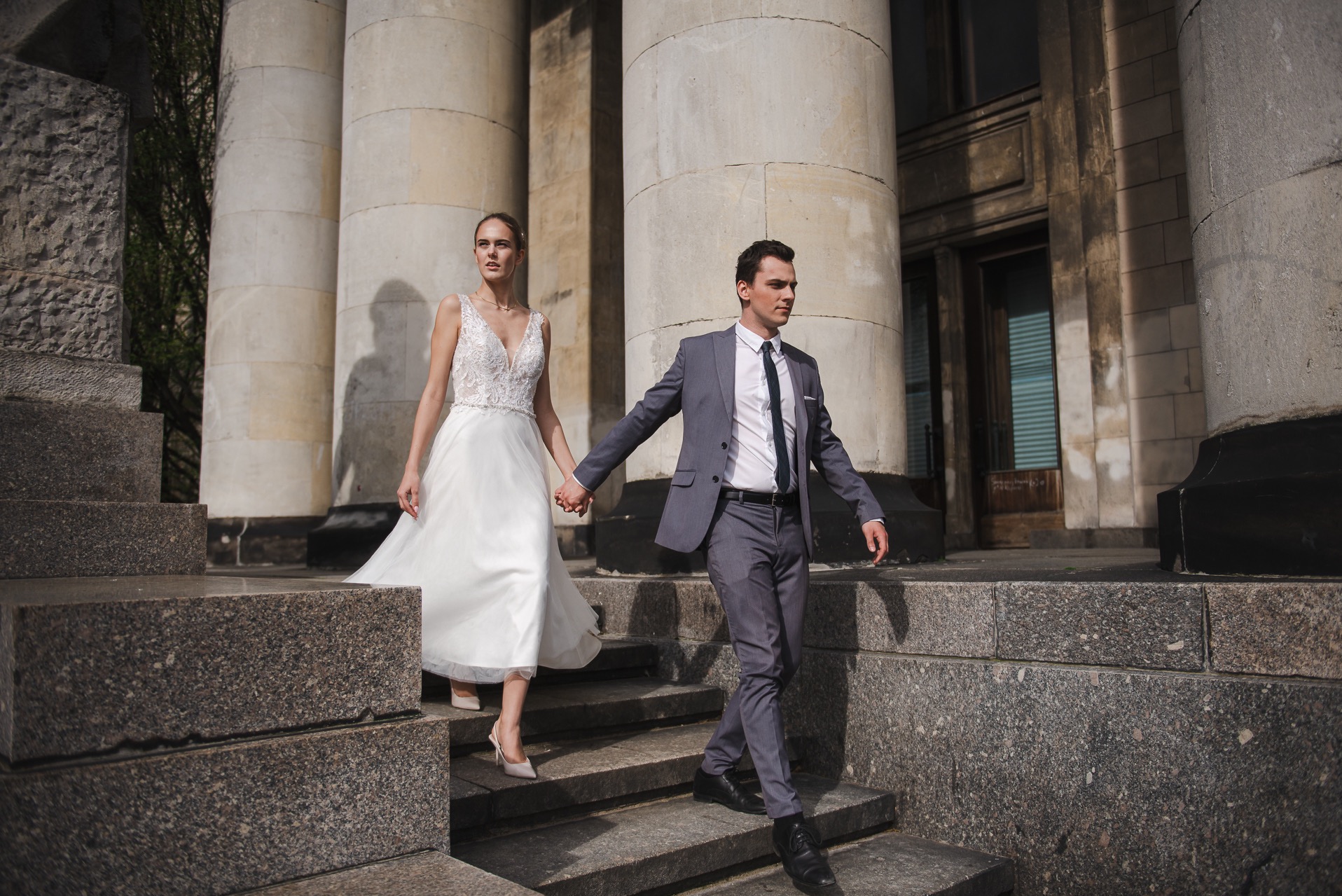 Para młoda schodzi po kamiennych schodach na tle kolumn. Panna młoda w koronkowej sukni, pan młody w szarym garniturze. Ujęcie z dołu, podkreślające architekturę.