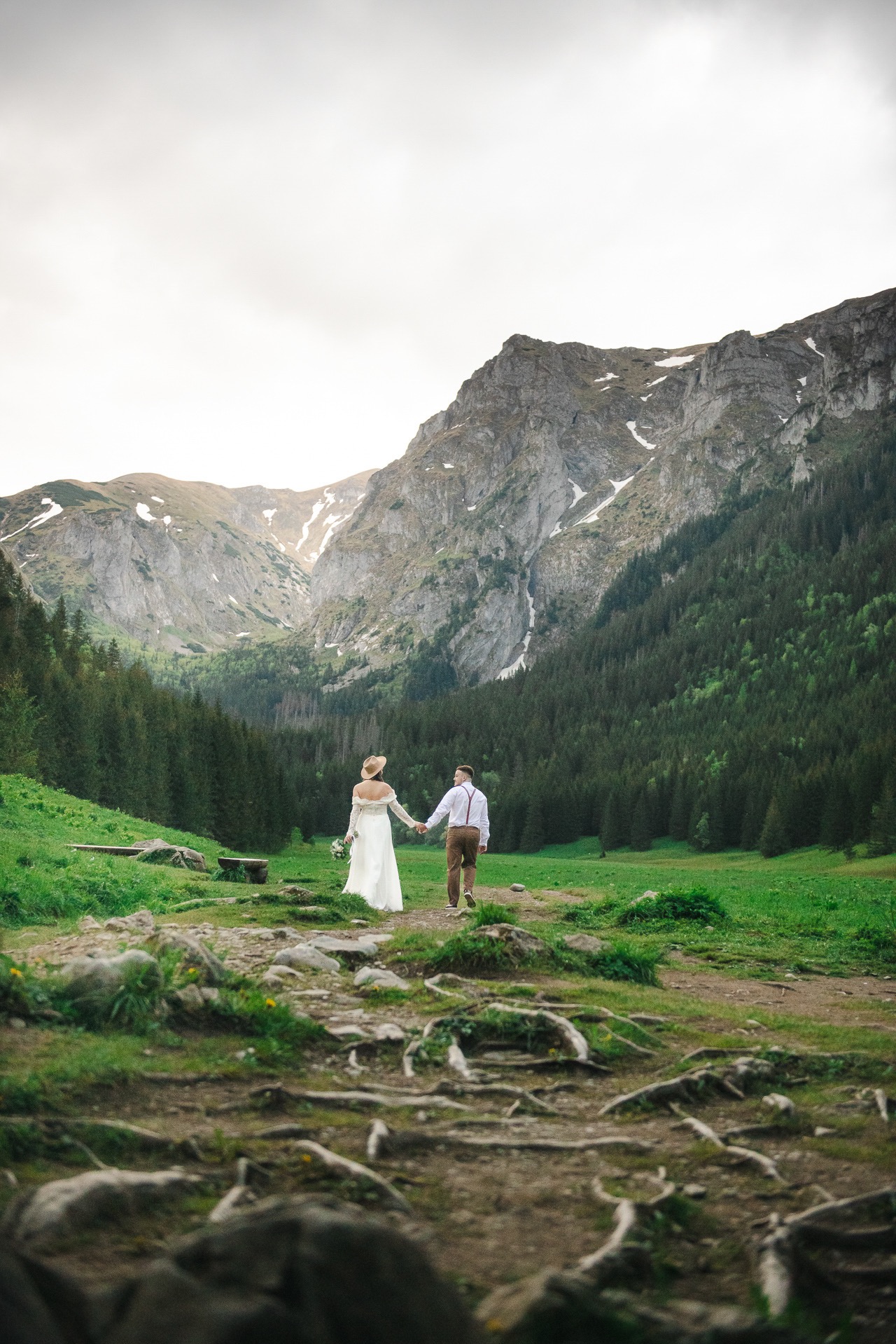 Para młoda, trzymając się za ręce, spaceruje w kierunku gór. Ona w białej sukni i kapeluszu, on w brązowych spodniach i białej koszuli z szelkami. W tle las i góry.