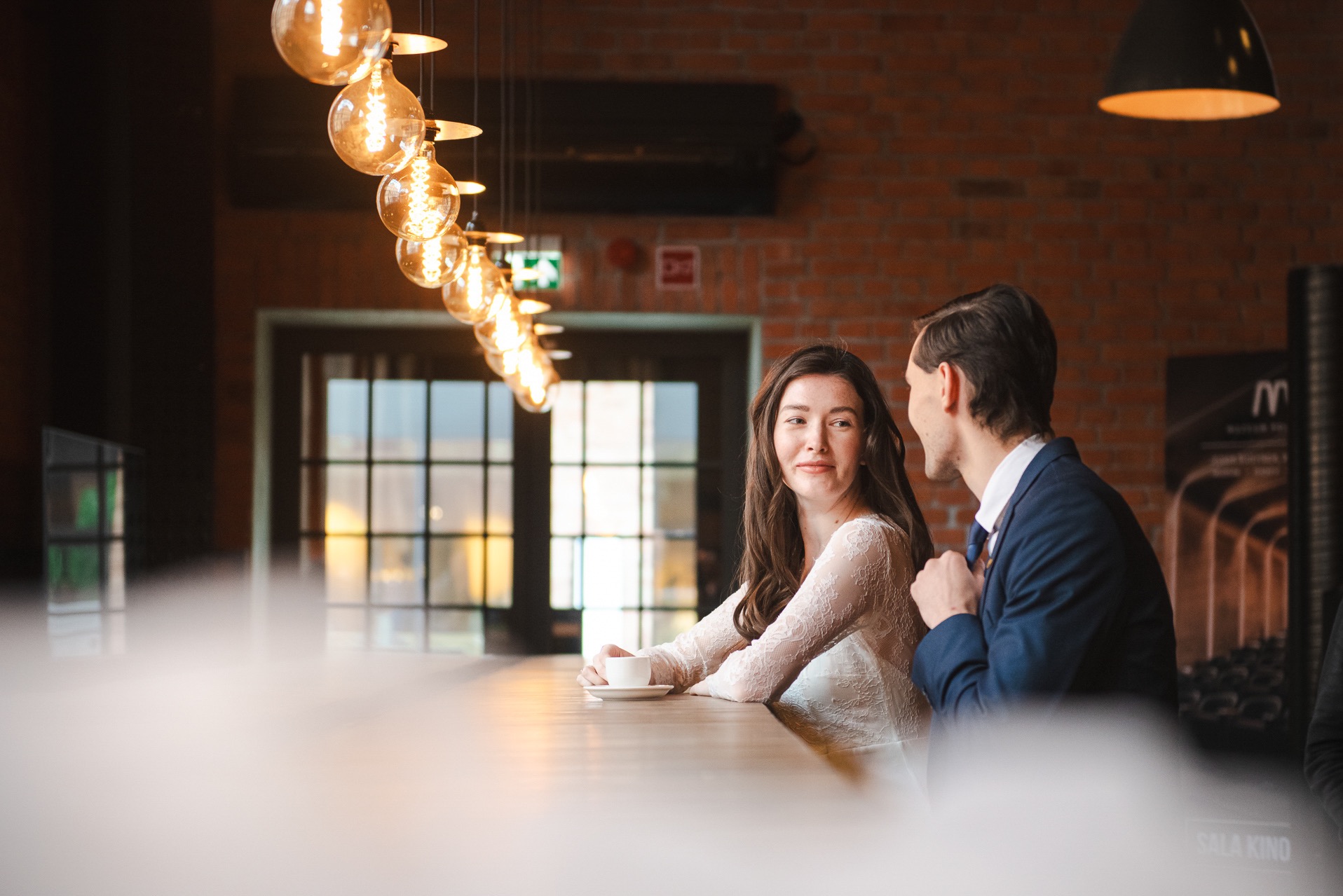 Uśmiechnięta panna młoda w koronkowej sukni siedzi przy barze z panem młodym w granatowym garniturze, pod dekoracyjnymi żarówkami w ciepłym świetle.