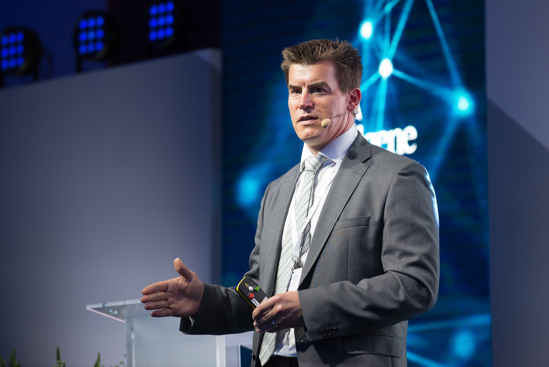 Mężczyzna w szarej marynarce i krawacie, z mikrofonem, przemawia na tle podświetlonej sceny z teleprompterem. W ręku trzyma urządzenie sterujące prezentacją.