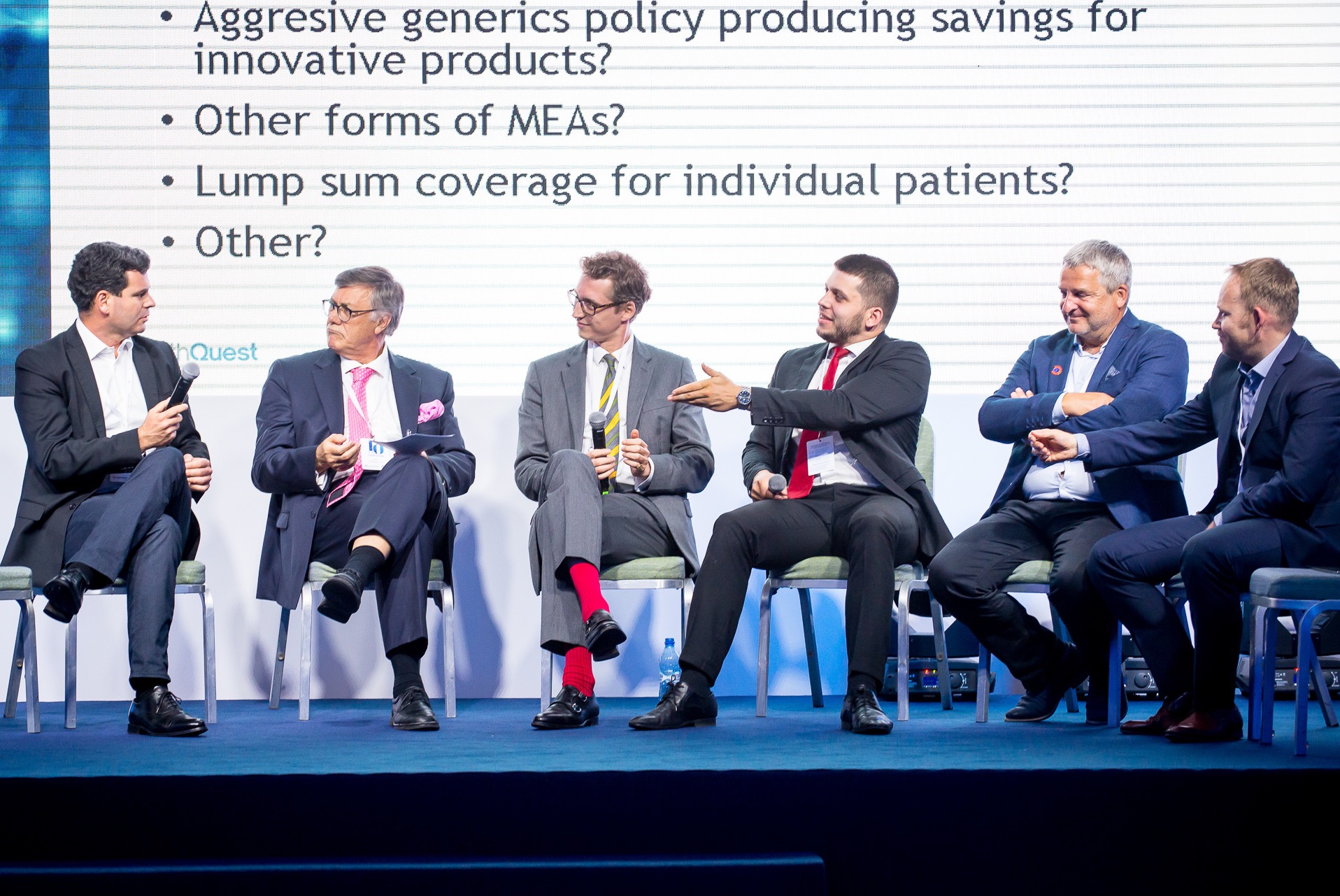 Panel dyskusyjny z sześcioma mężczyznami w garniturach siedzącymi na krzesłach na scenie. Jeden z nich gestykuluje ręką, a w tle widoczny ekran z pytaniami.