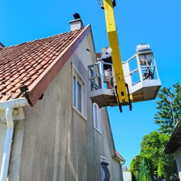LUK USŁUGI PODNOŚNIKOWE - Pracownik w podnośniku koszowym czyści elewację domu. Widoczne ślady mycia na ścianie, okna z białymi ramami i czerwony dach. Błękitne niebo w tle.