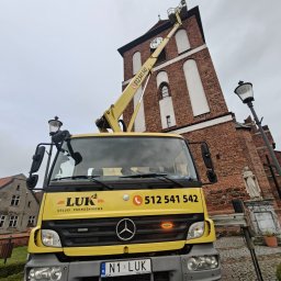 LUK USŁUGI PODNOŚNIKOWE - Żółty podnośnik koszowy LUK na tle ceglanej wieży kościoła z zegarem. Kosz podnośnika na wysokości zegara. Widoczny numer telefonu i logo firmy.