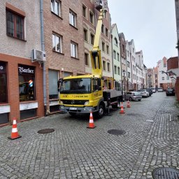 LUK USŁUGI PODNOŚNIKOWE - Żółty podnośnik koszowy LUK na brukowanej ulicy, z wysięgnikiem sięgającym do okna kamienicy. Pomarańczowe pachołki zabezpieczają obszar robót. Widok na starą zabudowę miejską.