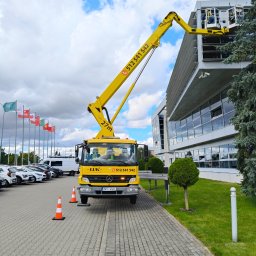 LUK USŁUGI PODNOŚNIKOWE - Żółty podnośnik koszowy LUK na chodniku, z pracownikami w koszu przy szklanej fasadzie budynku. Flagi w tle, oznakowanie stożkami.
