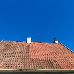 LUK USŁUGI PODNOŚNIKOWE - Czerwona dachówka z anteną i kominami na tle błękitnego nieba. Widoczne ślady zabrudzeń na dachu. Ujęcie z dołu, podkreślające strukturę i kolor dachu.