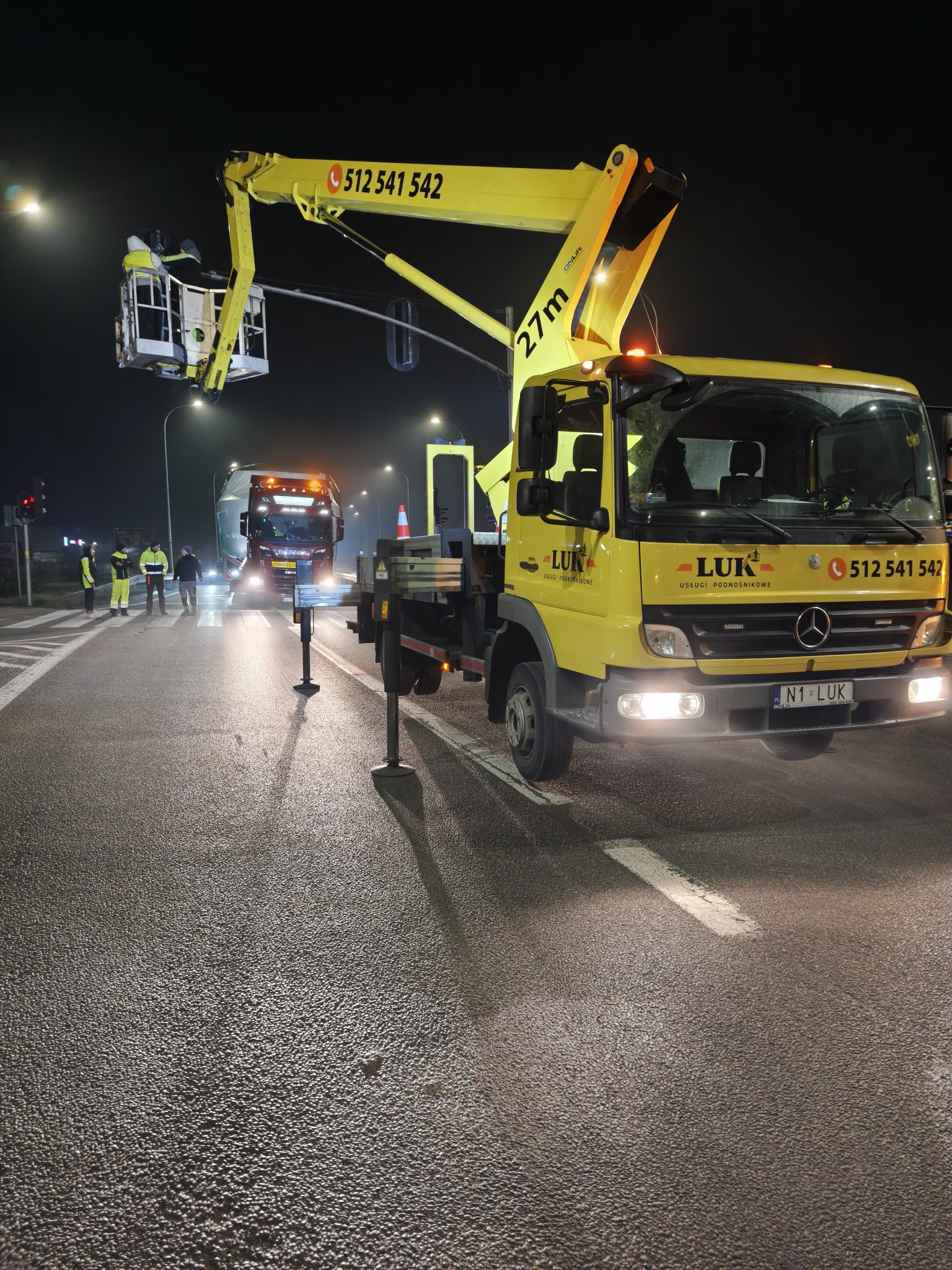 Żółty podnośnik koszowy LUK na drodze nocą, z wysięgnikiem ustawionym nad jezdnią. Widoczni pracownicy w odblaskowych ubraniach i nadjeżdżająca ciężarówka.