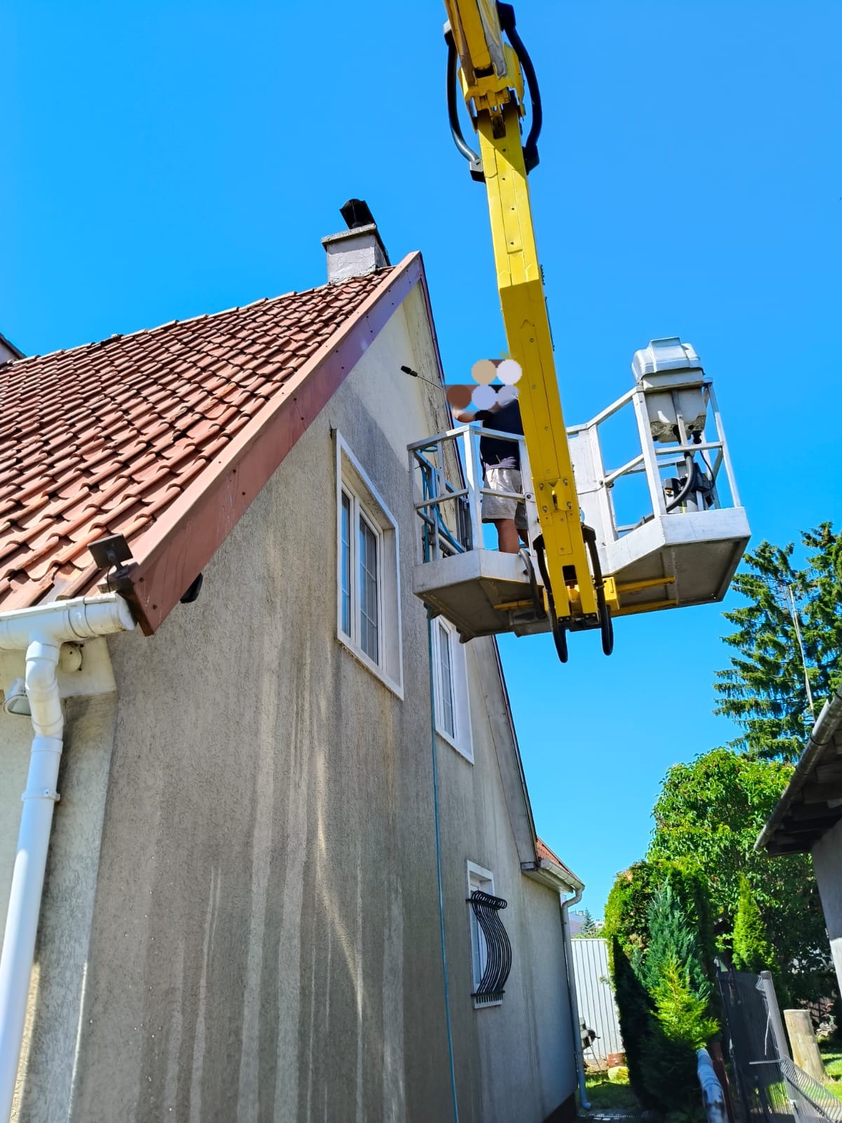 Pracownik w podnośniku koszowym czyści elewację domu. Widoczne ślady mycia na ścianie, okna z białymi ramami i czerwony dach. Błękitne niebo w tle.