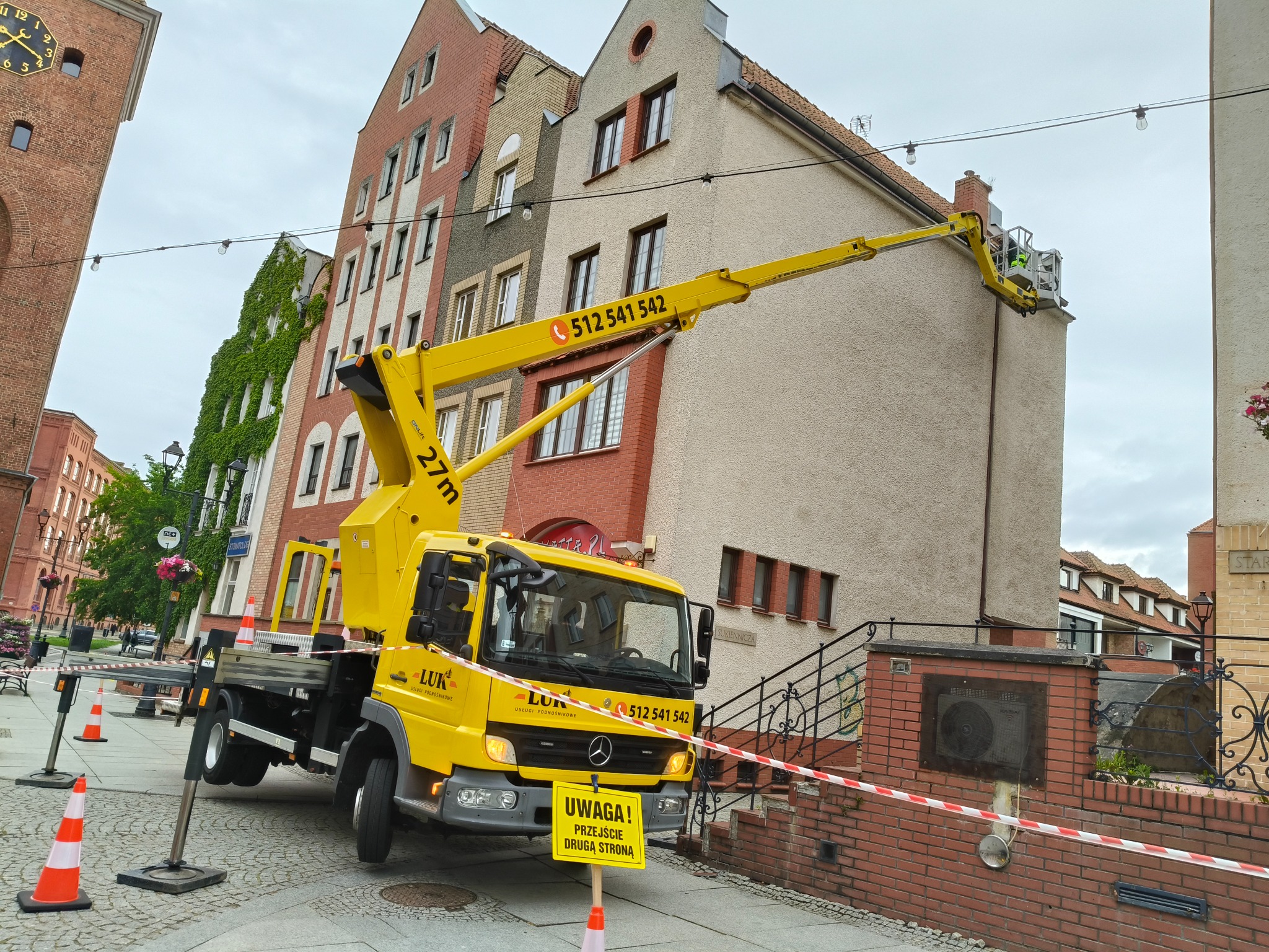 Żółty podnośnik koszowy LUK na tle kamienic podczas prac wysokościowych. Na wysięgniku pracownik w kasku. Ulica zabezpieczona taśmą i pachołkami.
