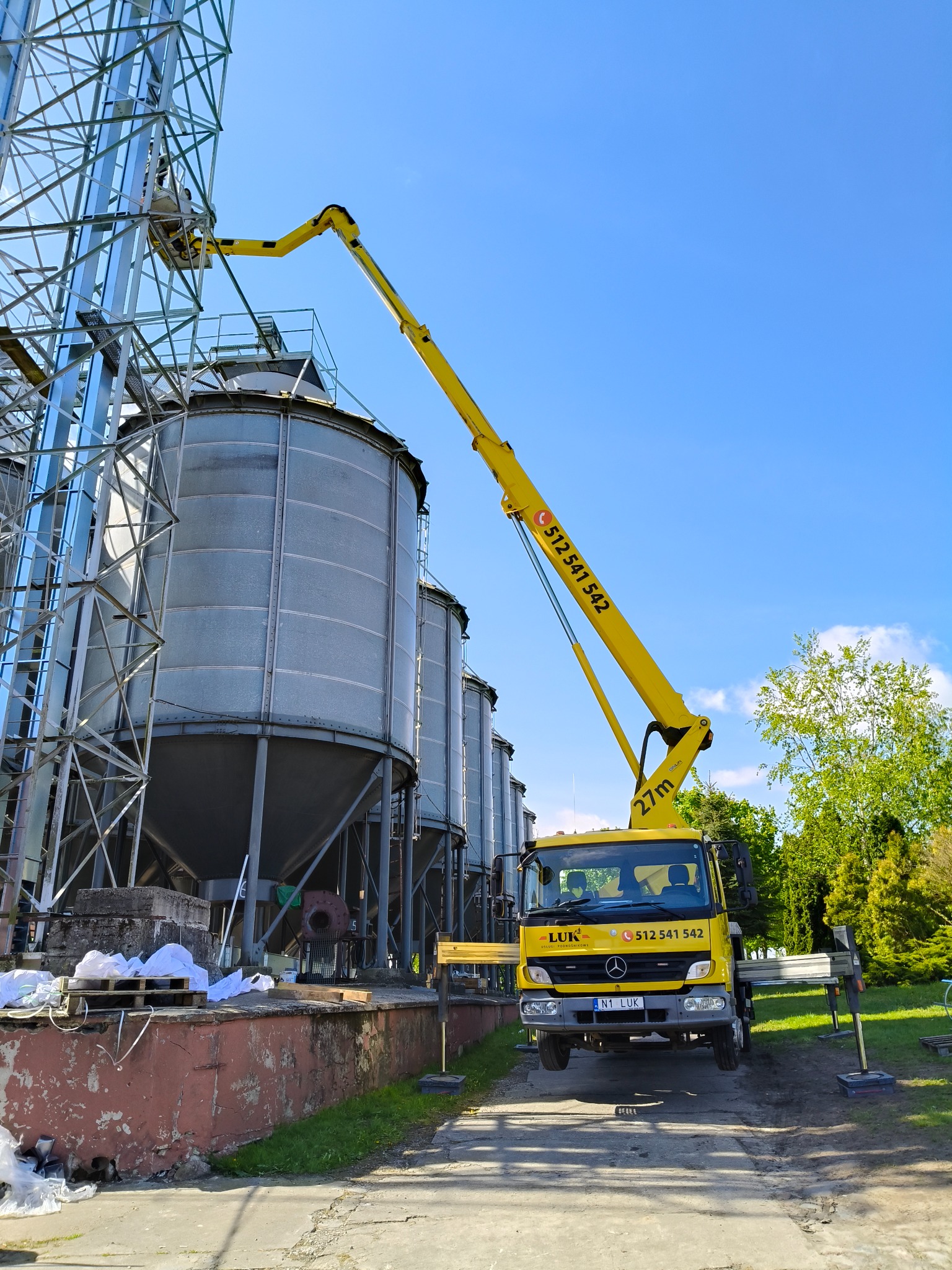 Żółty podnośnik koszowy LUK na tle silosów zbożowych i błękitnego nieba. Pracownik w koszu wykonuje prace na wysokości. Widoczne podpory stabilizujące pojazd.